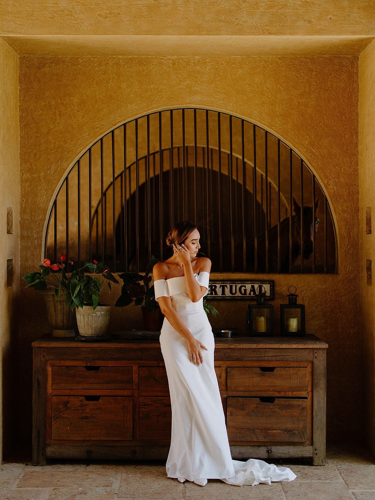 Bride in elegant white off-shoulder gown poses against rustic arch and wooden furniture.