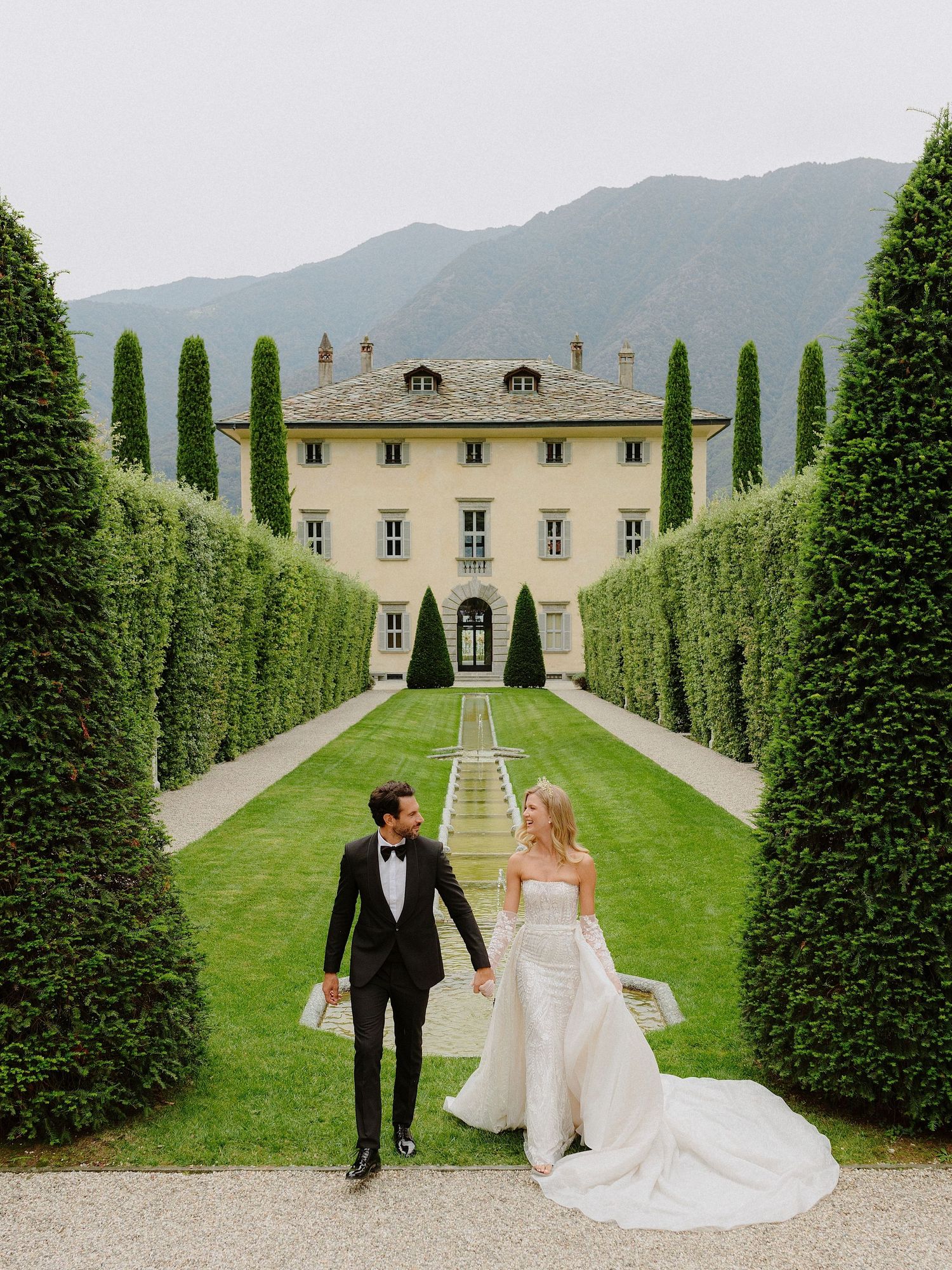 Couple walking hand in hand on formal garden path leading to Italian villa.