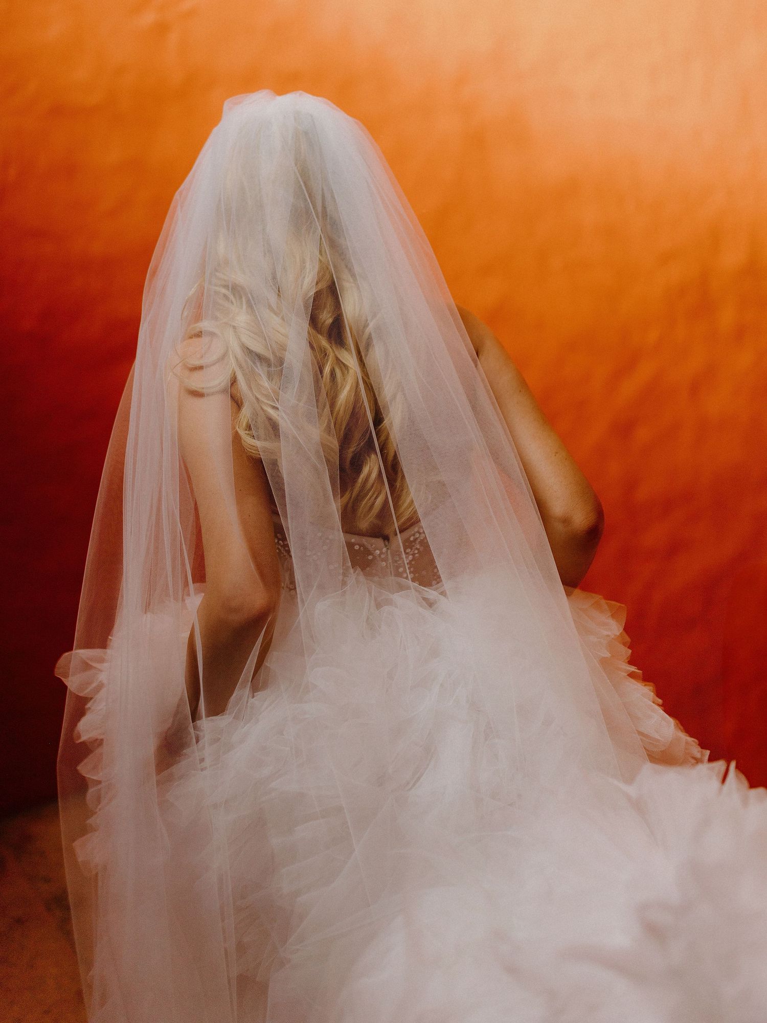 Artistic bridal portrait with flowing veil against vibrant orange background.