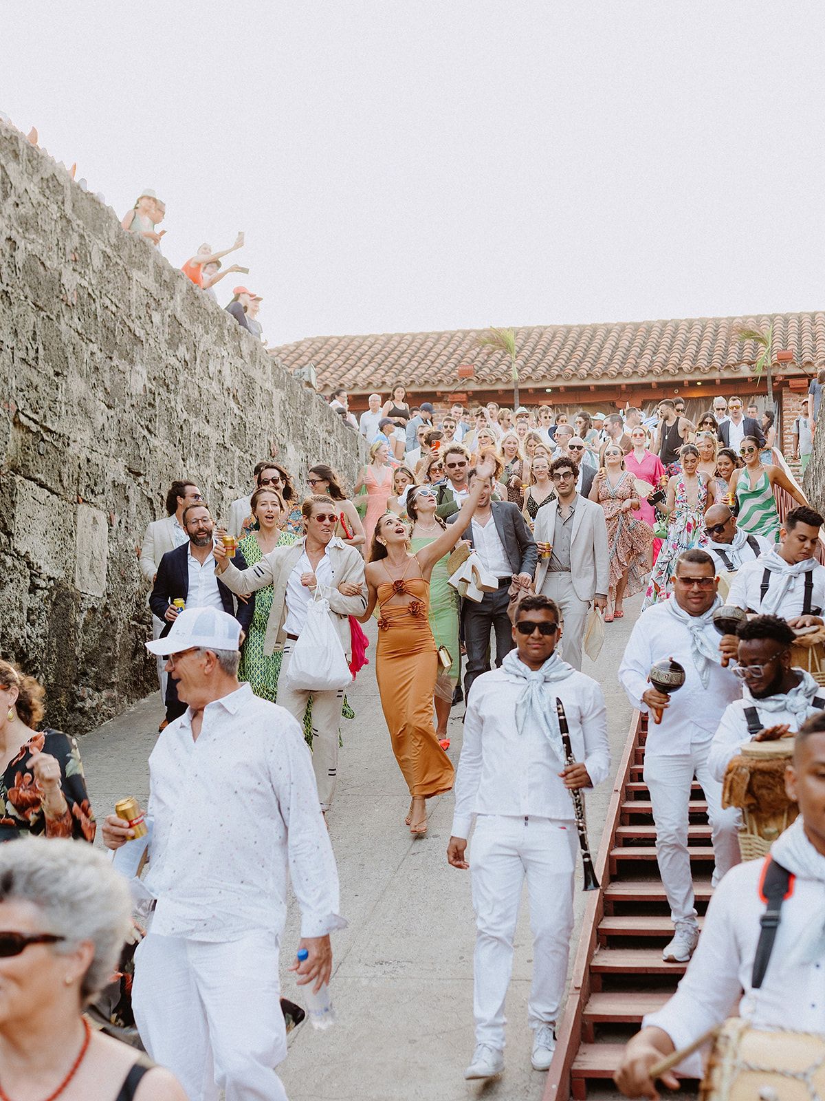 Wedding party in white attire descending stone steps during celebration.