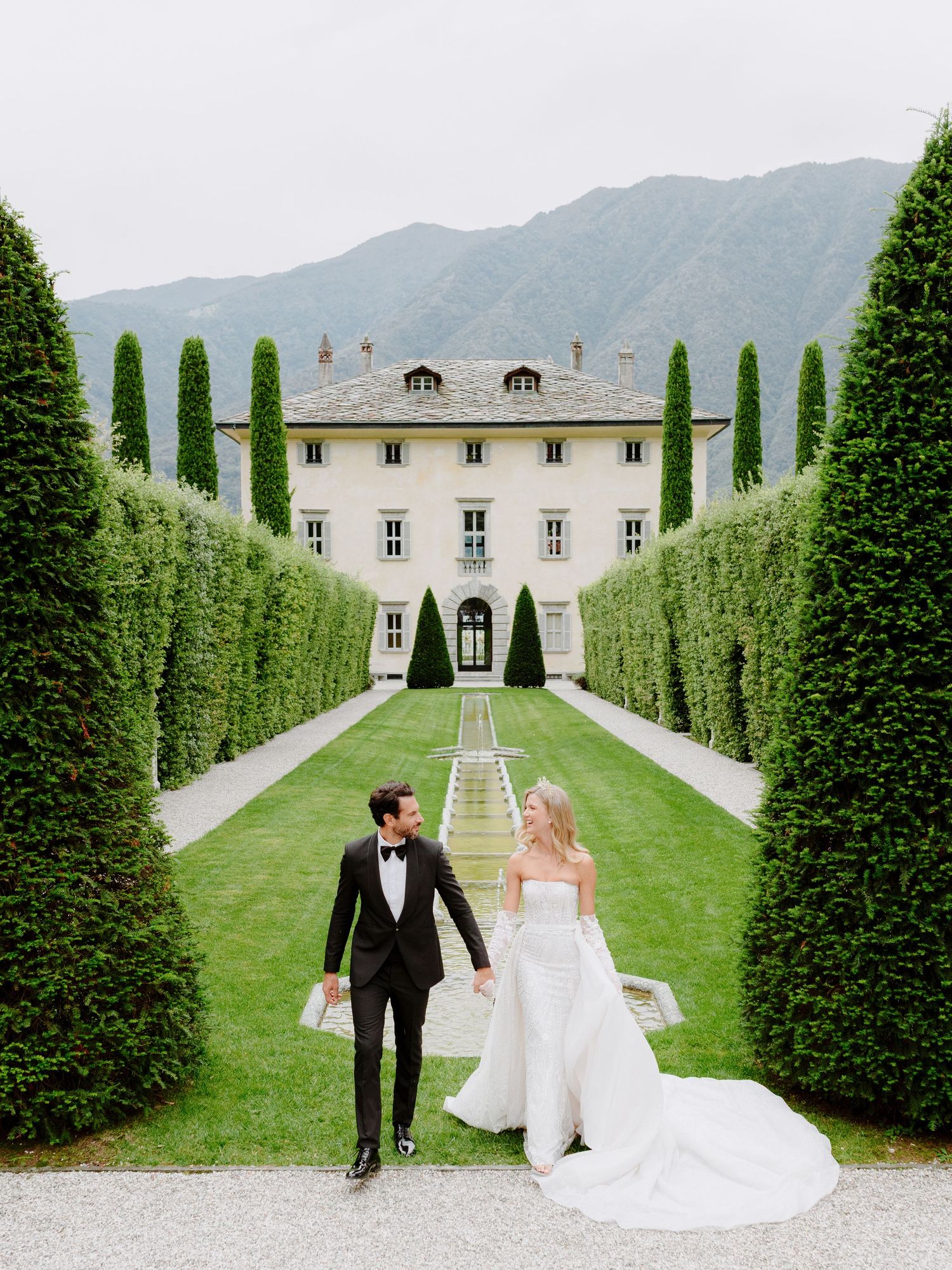 A majestic villa with symmetrical cypress trees and manicured lawn leads to a grand mansion in the Italian mountains.