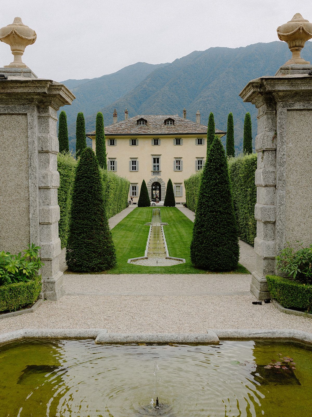 Elegant villa with manicured cypress gardens and formal landscaping against mountain backdrop in Lake Como.