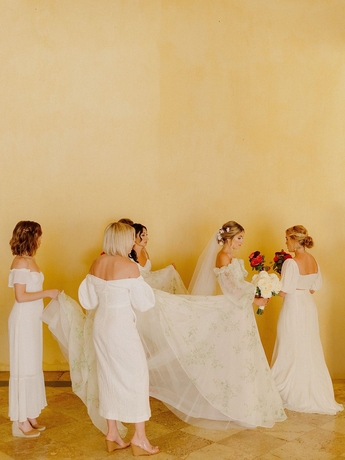 Bridesmaids in white dresses helping arrange a wedding veil against a golden wall.