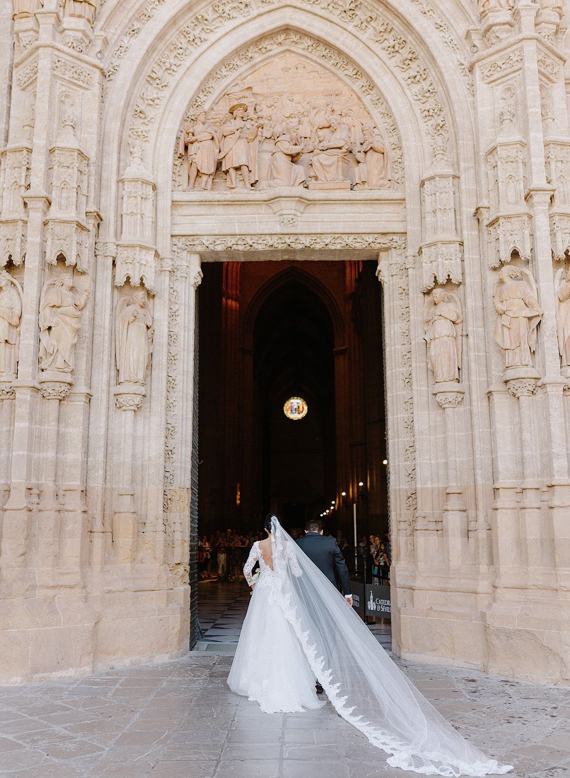 A long wedding veil trails behind as a couple enters an ornate cathedral with detailed stone carvings.