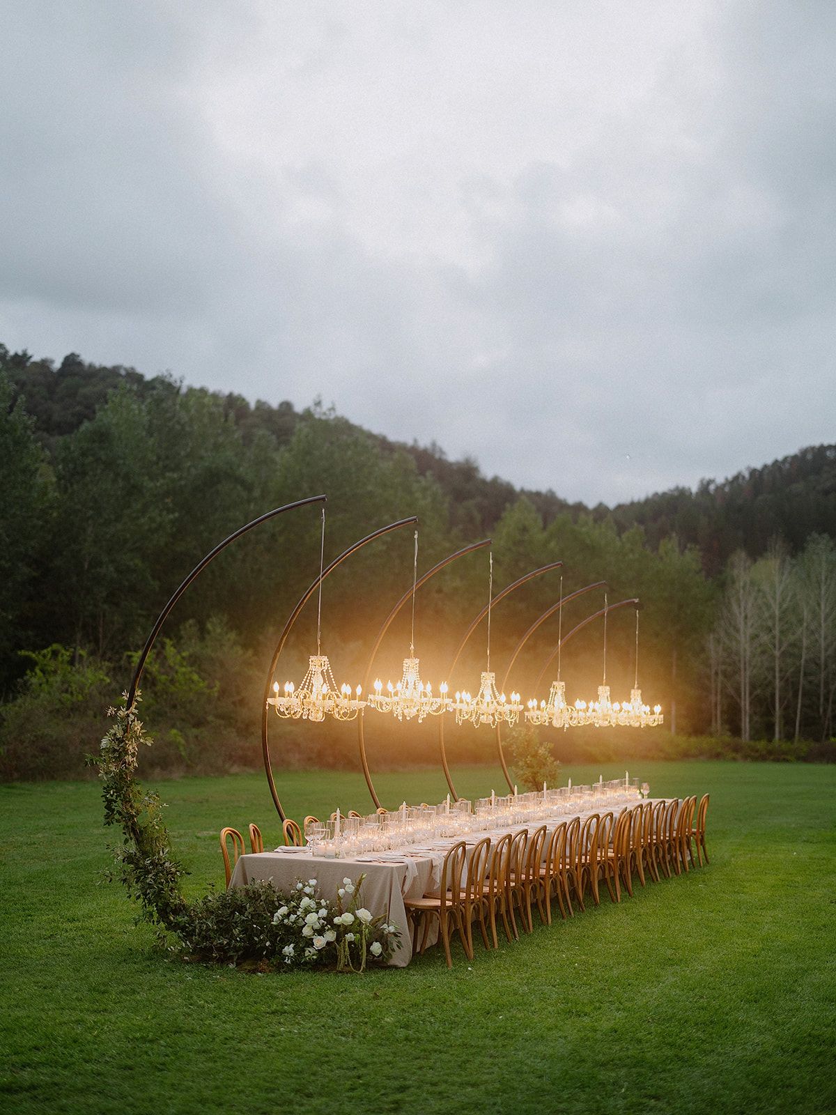 Elegant outdoor wedding reception table with hanging chandeliers and floral arches in a mountain setting.