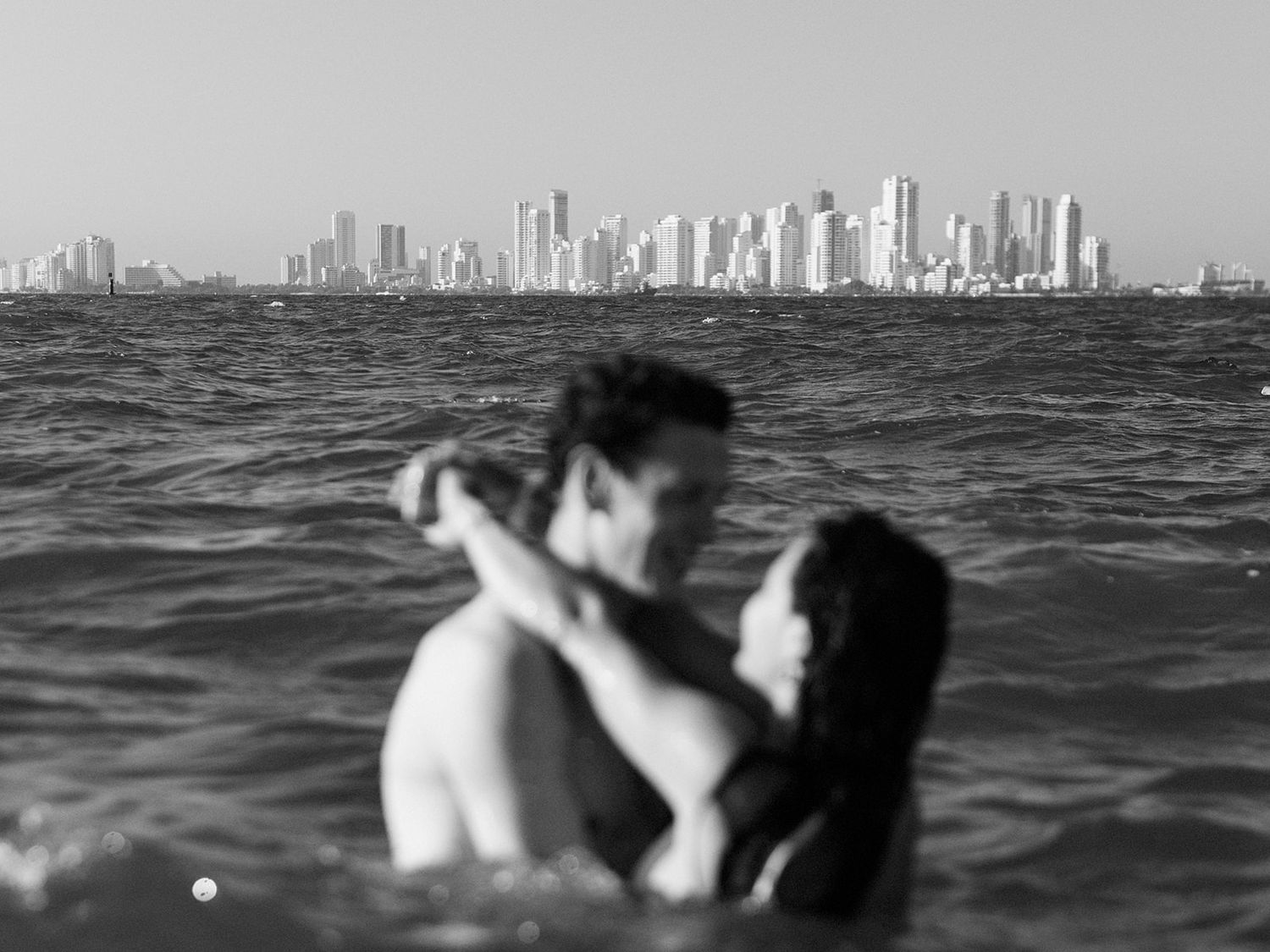 Romantic couple embracing in ocean waters with city skyline visible in background.