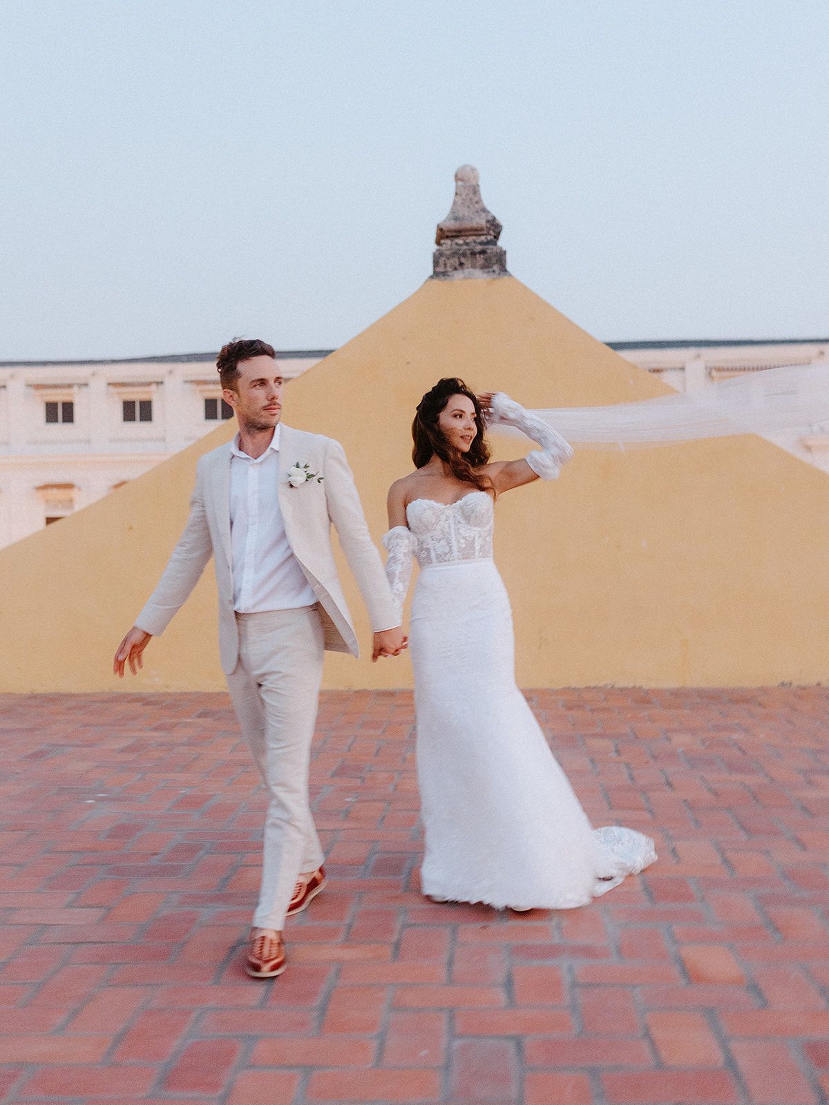Newlyweds walking hand in hand on terracotta tiles with colonial architecture in background.