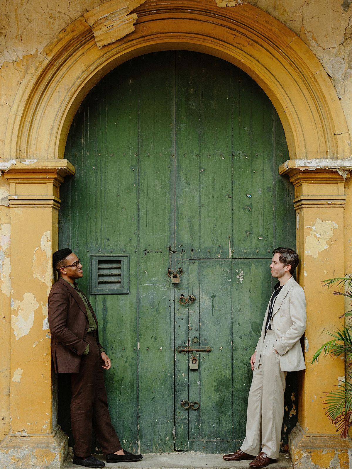 Two people posing by a weathered green colonial door with yellow archway.