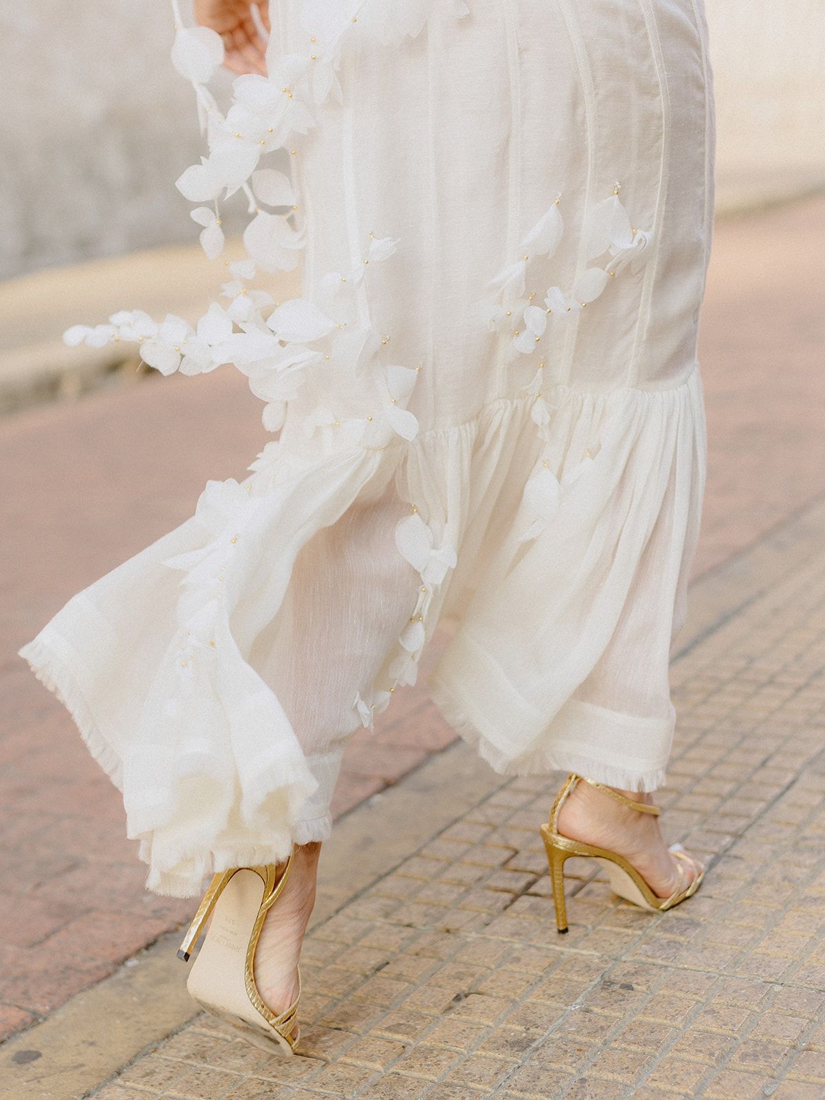 Close-up of flowing white wedding dress hem and gold high heels on cobblestone street.