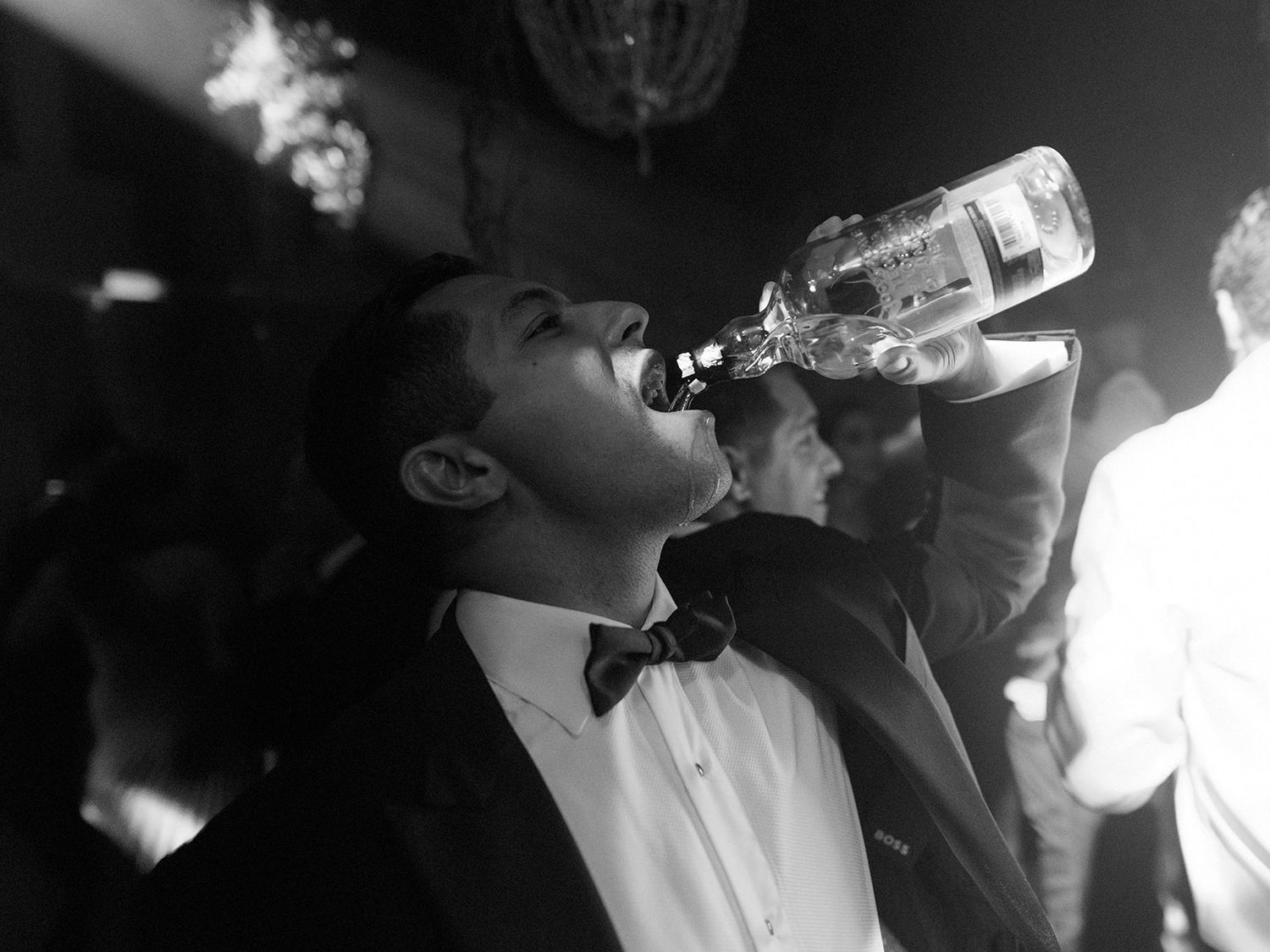 Black and white photo of someone in a tuxedo drinking from a bottle at a celebration.