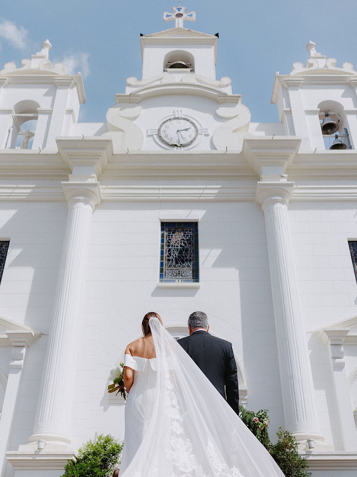 Bride and father walking towards white church entrance with dramatic flowing veil.