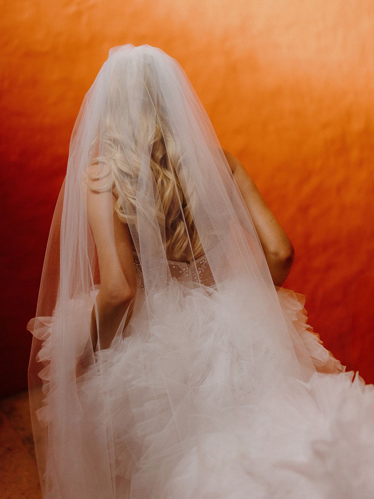 Ethereal bridal portrait with flowing veil against vibrant orange background.