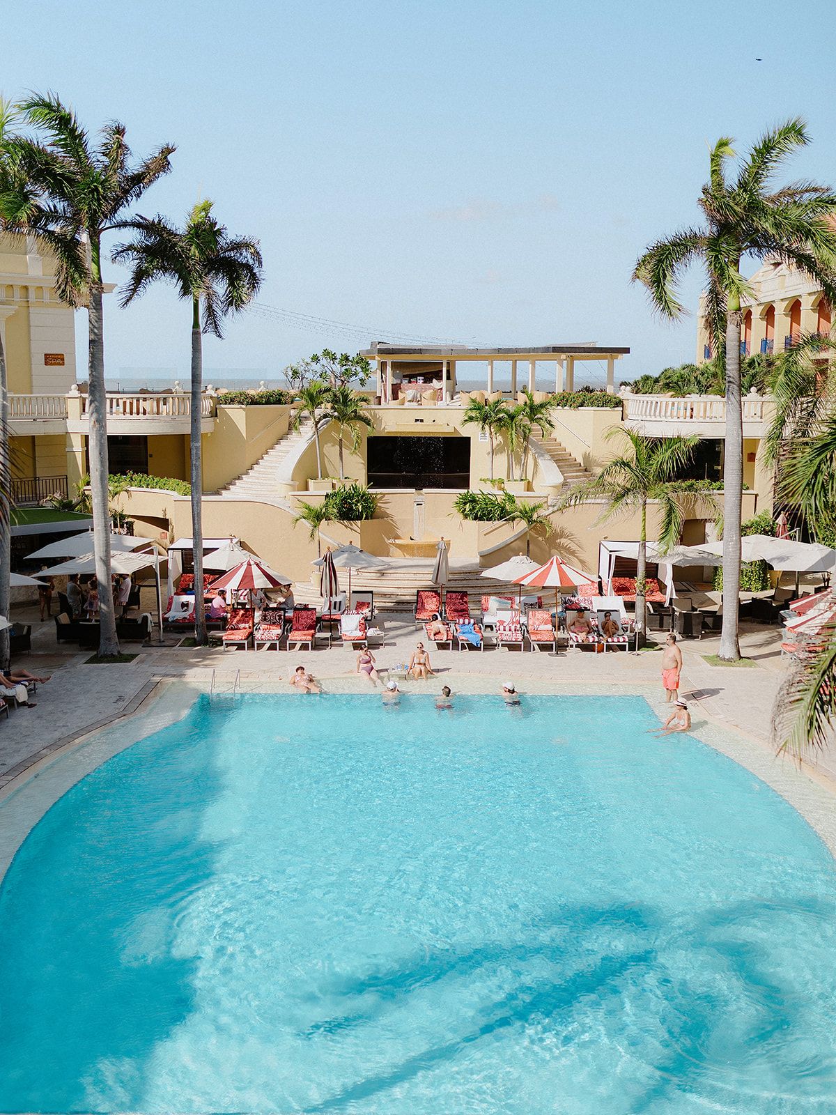 Resort swimming pool surrounded by palm trees and umbrellas in a tropical setting.
