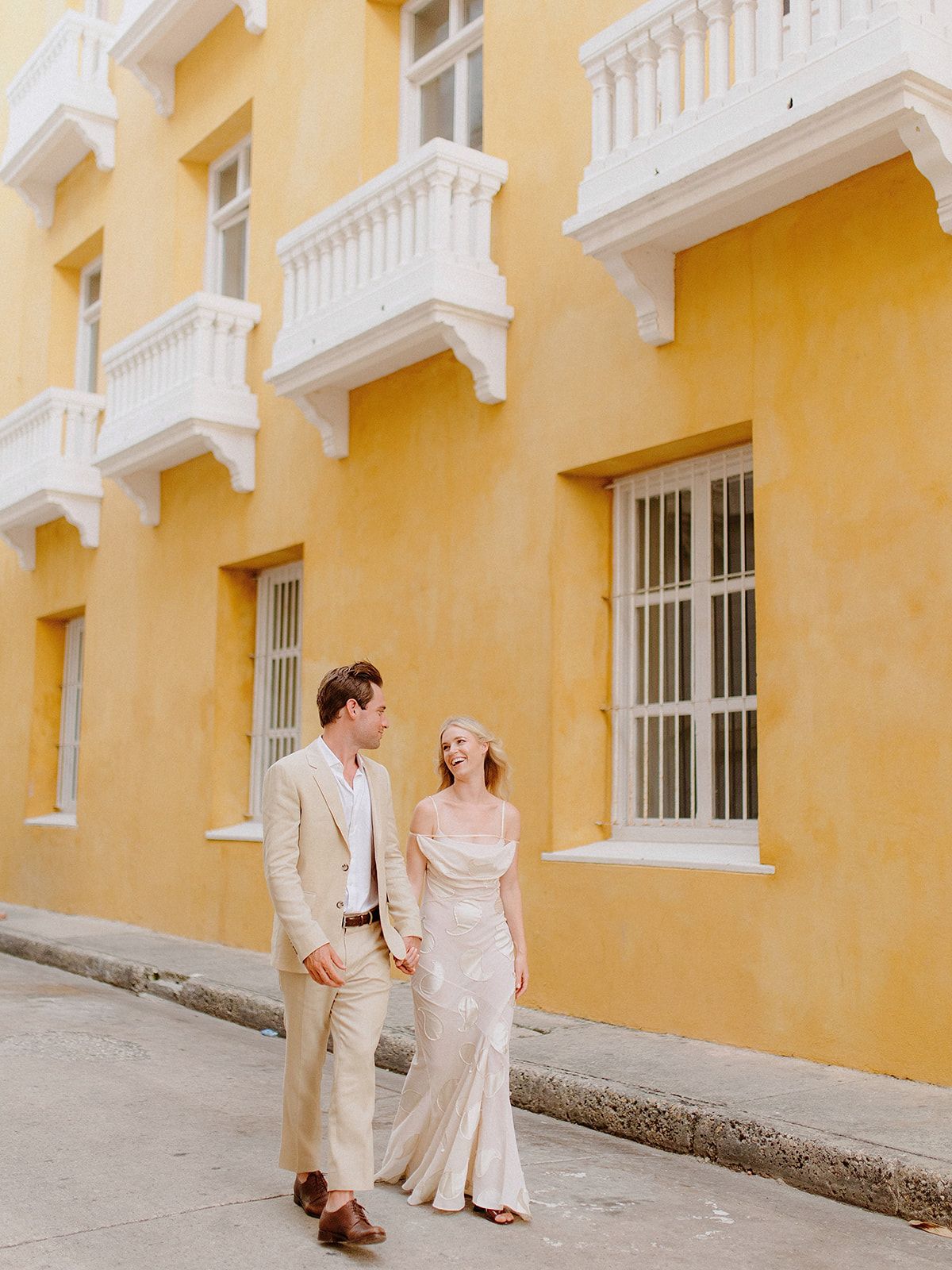 A couple walking along a yellow colonial building with white balconies in a tropical setting.
