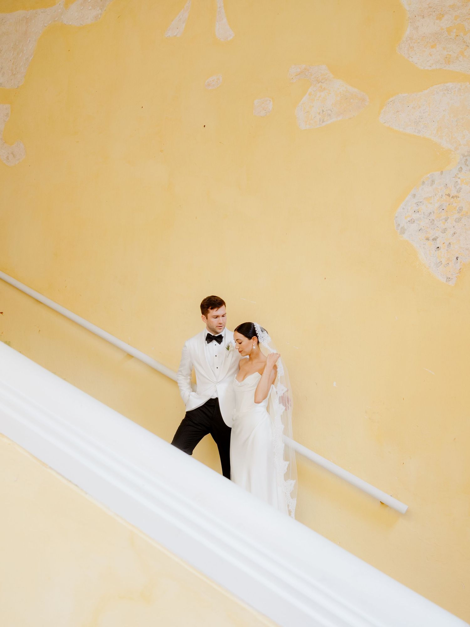 A bride and groom stand together on white steps against a yellow wall during their wedding photoshoot.
