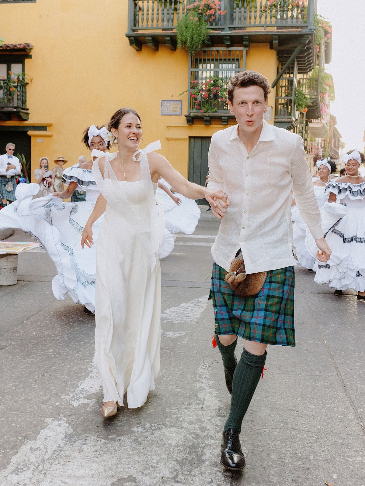 Wedding celebration in colorful street with traditional dancers and couple in Scottish attire.