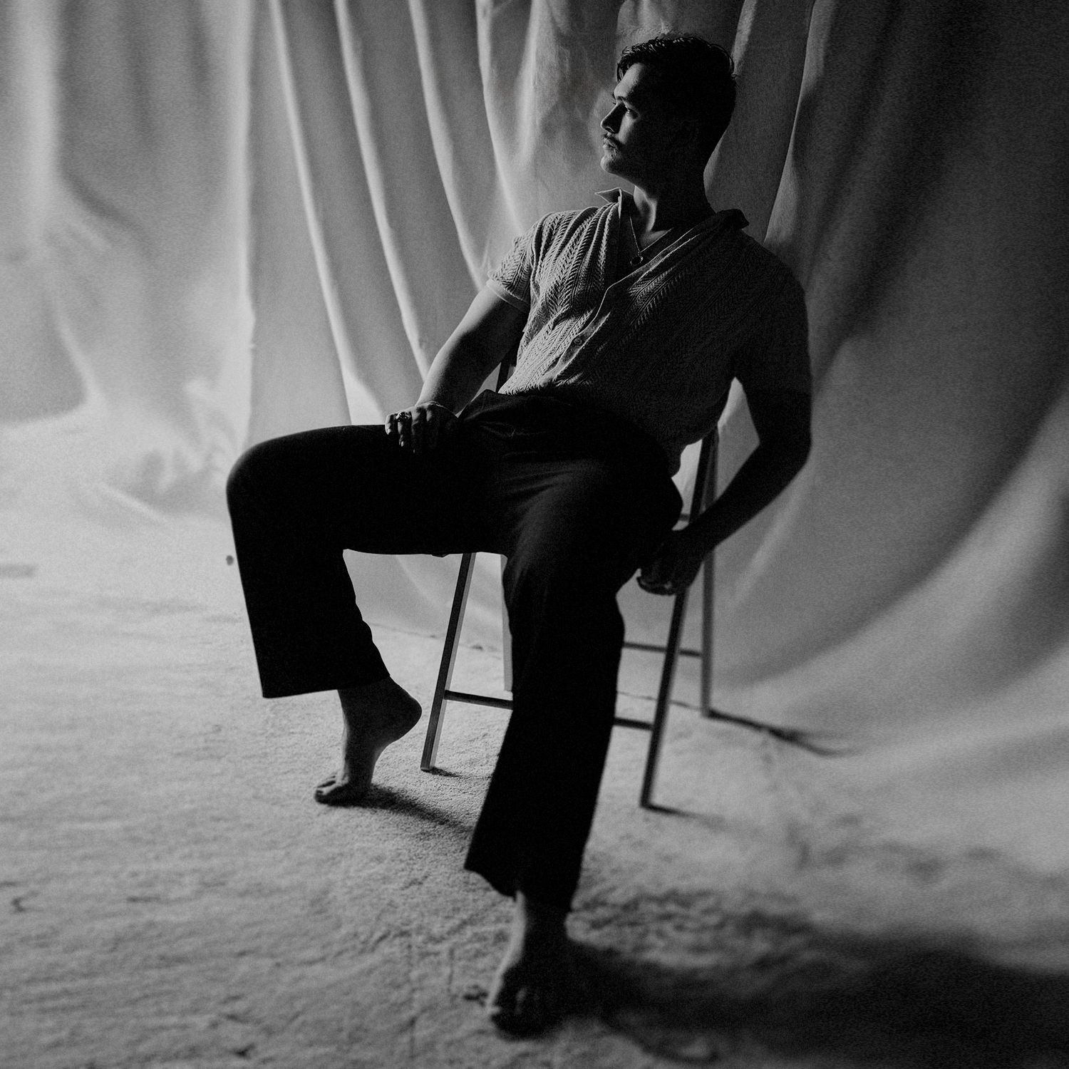 Artistic black and white silhouette of a figure seated on a chair against a draped backdrop.