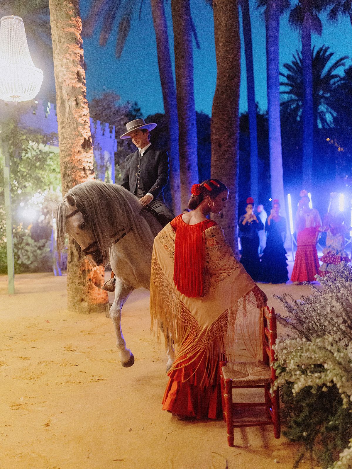 A festive evening scene with horseback riders and flamenco dancers among palm trees.