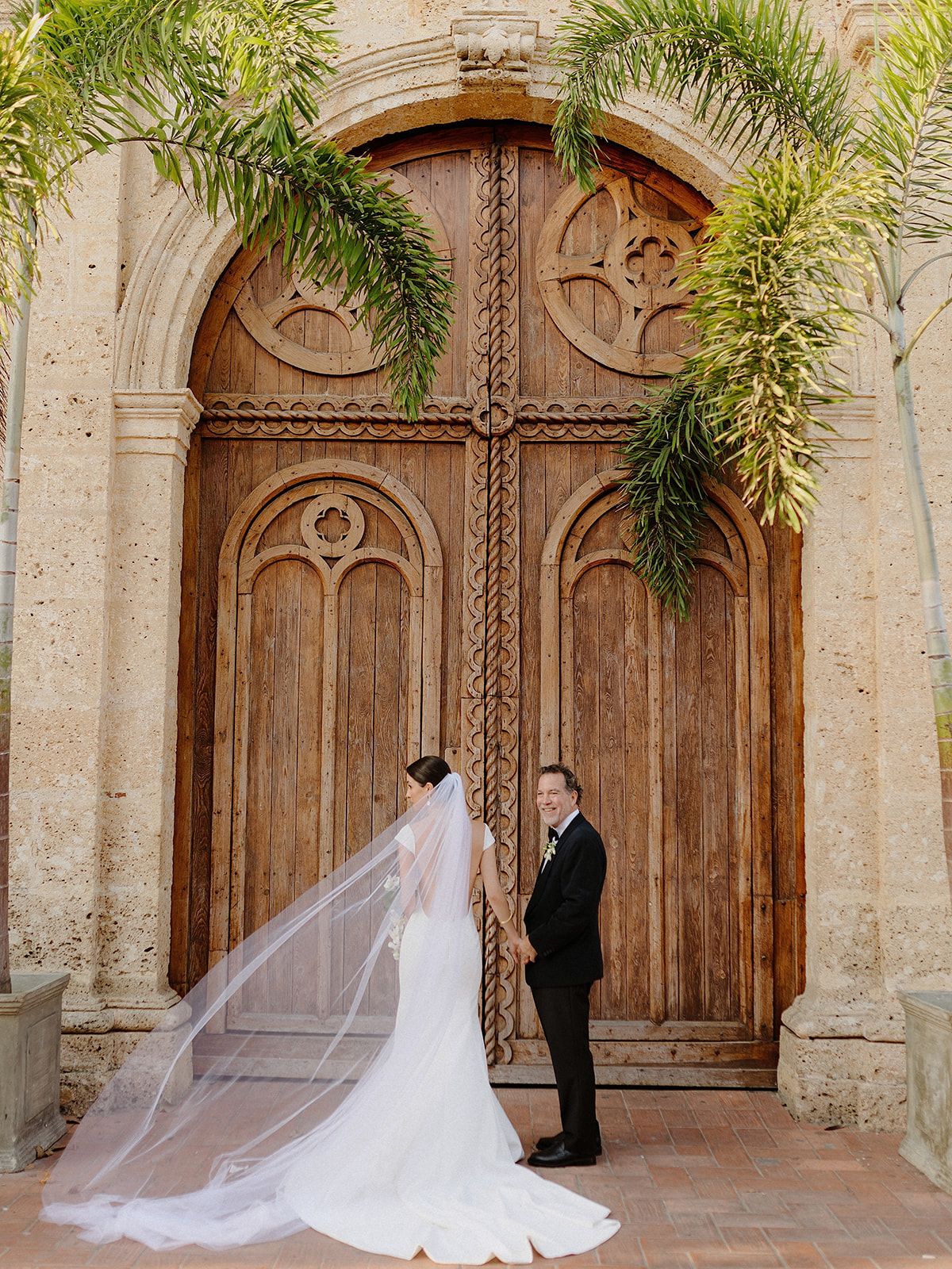 Classic wedding portrait in front of grand wooden church doors adorned with palm fronds.