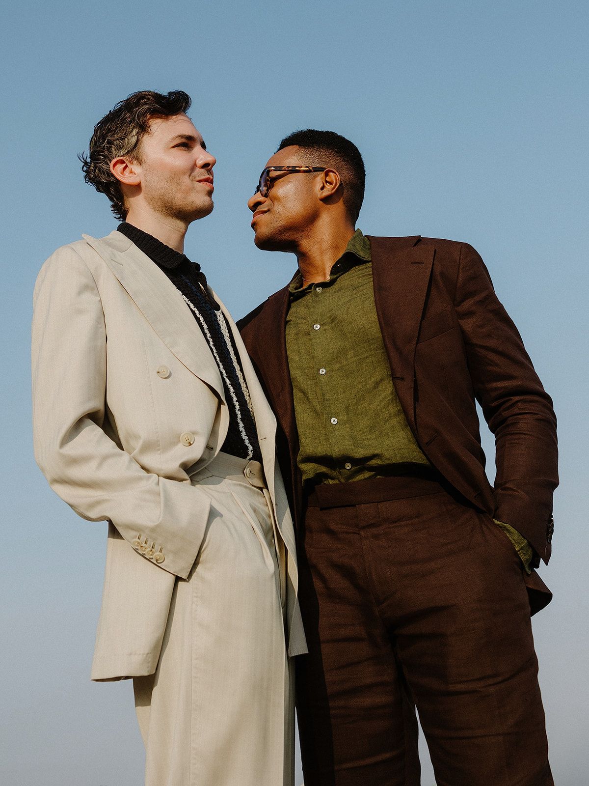 Two people in contrasting suits, one beige and one brown, pose together against a blue sky backdrop.