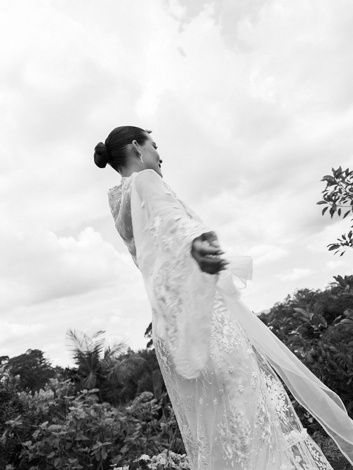 Dramatic black and white photo of bride in lace dress against cloudy sky.