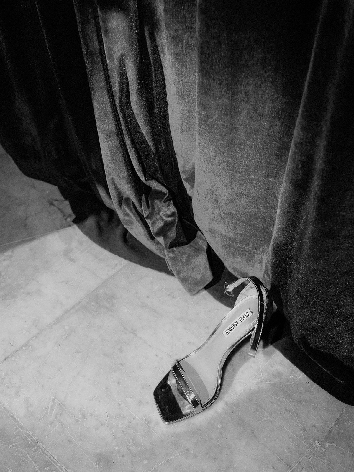 Black and white detail shot of elegant bridal shoe peeking out from beneath flowing dress.