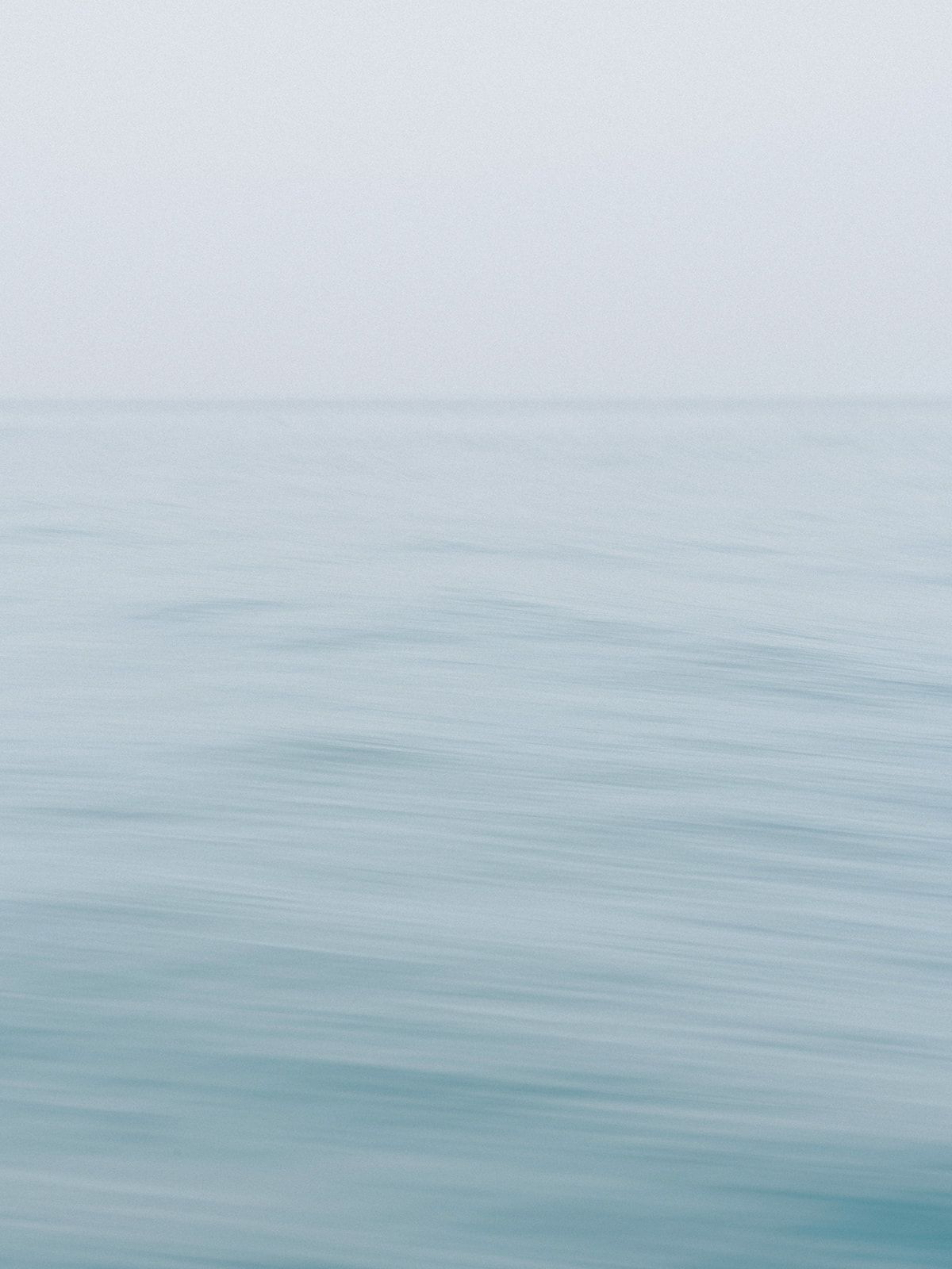 Minimalist seascape photograph with soft blue waters blending into misty horizon.