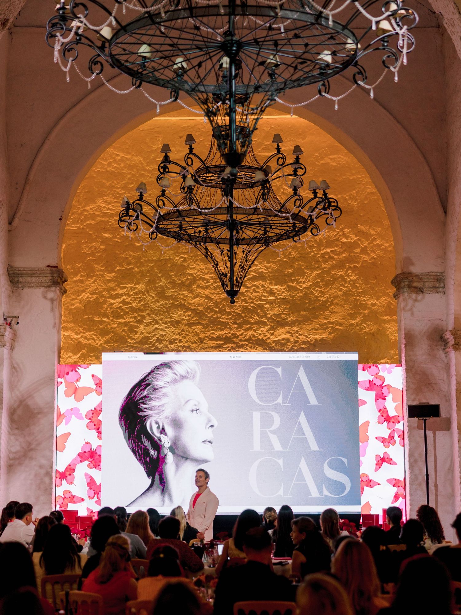 An ornate chandelier hangs above a presentation screen in an elegant venue with arched architecture and warm lighting.