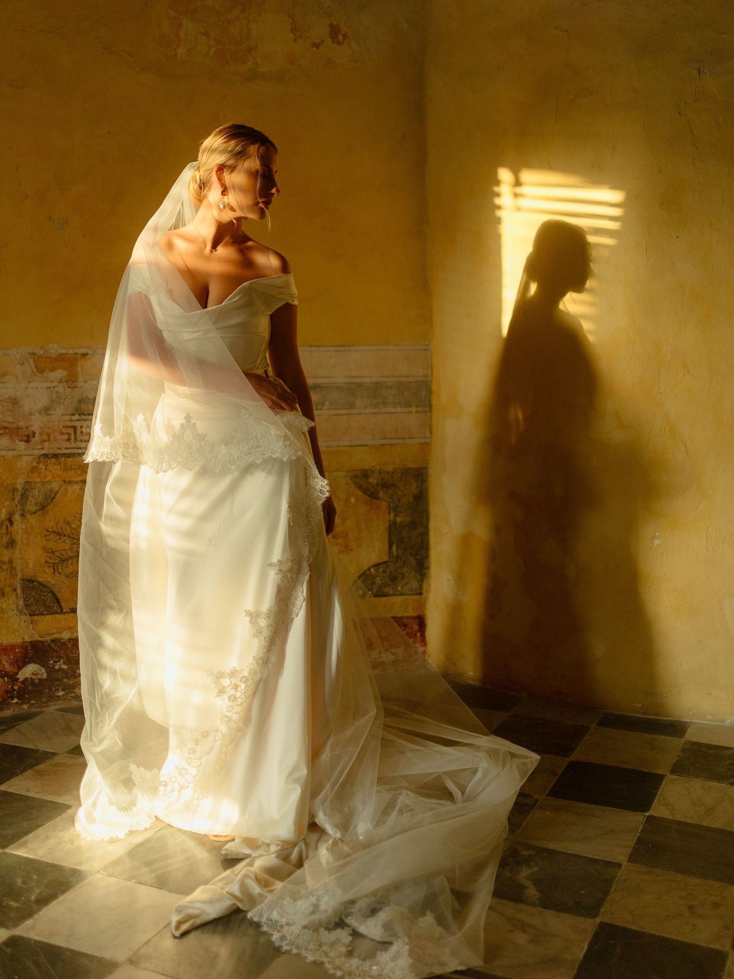A bride in a flowing white dress stands in golden sunlight casting a dramatic shadow on a weathered wall.