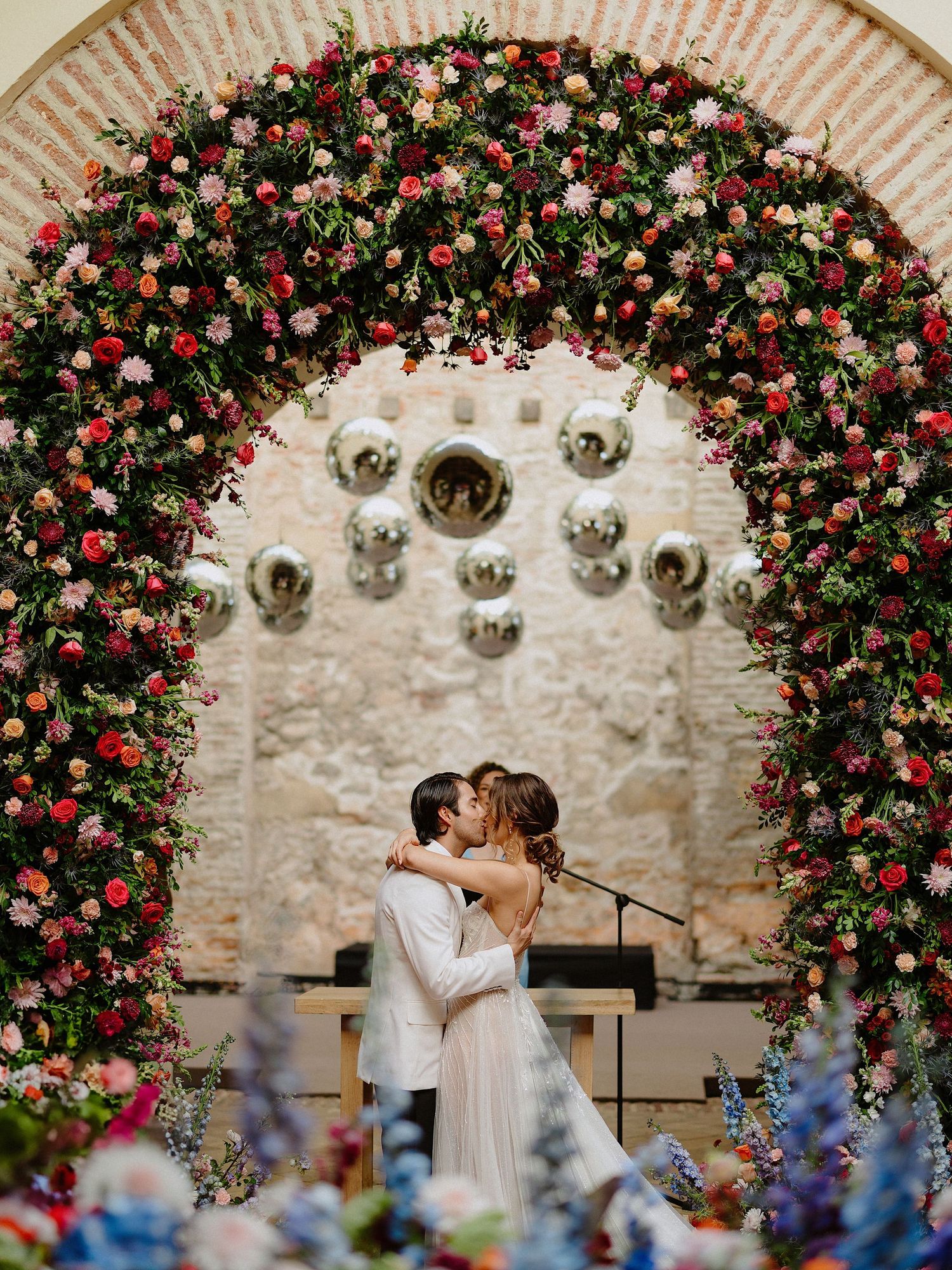 Wedding ceremony under a lush floral arch with pink and red roses and hanging lanterns.
