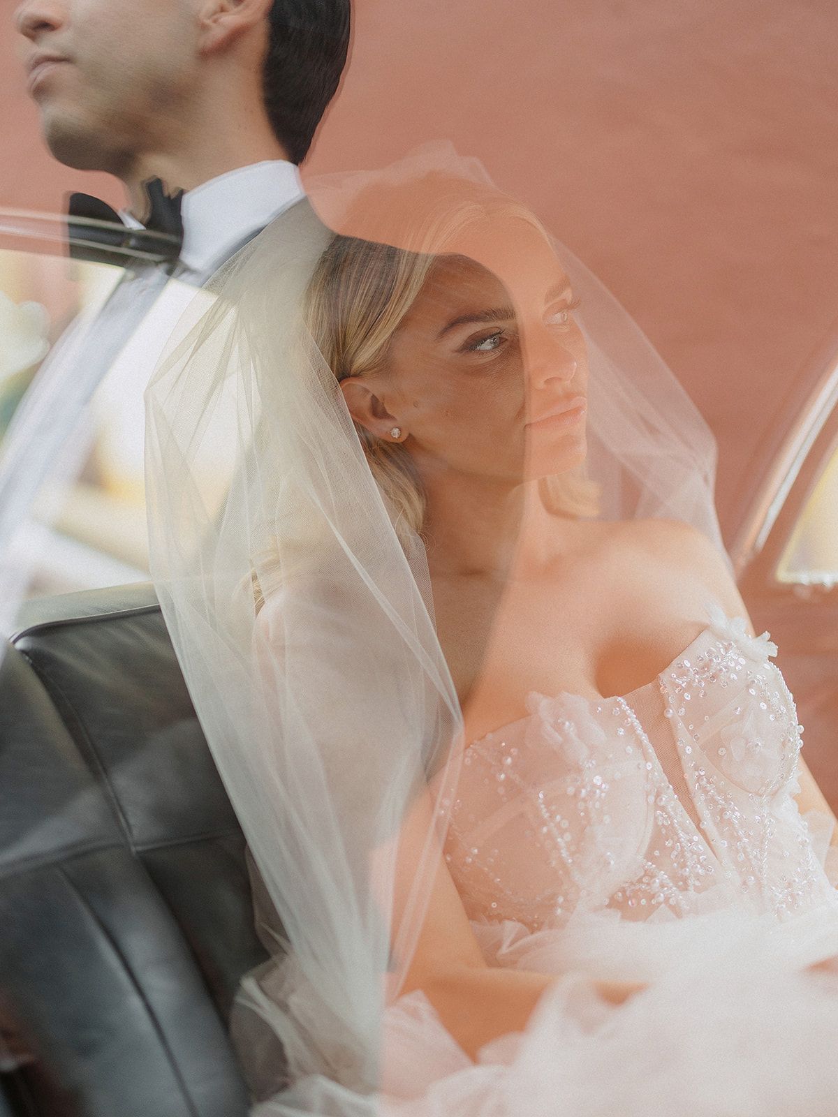 Bride in a delicate lace wedding dress and veil poses with groom in classic black tuxedo against coral backdrop.