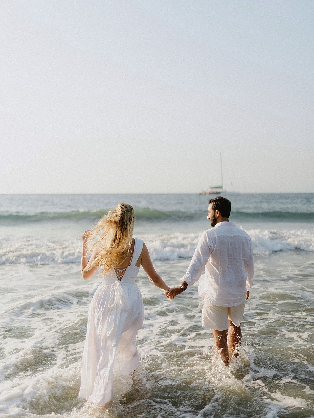 A couple holding hands while walking through ocean waves in white wedding attire.