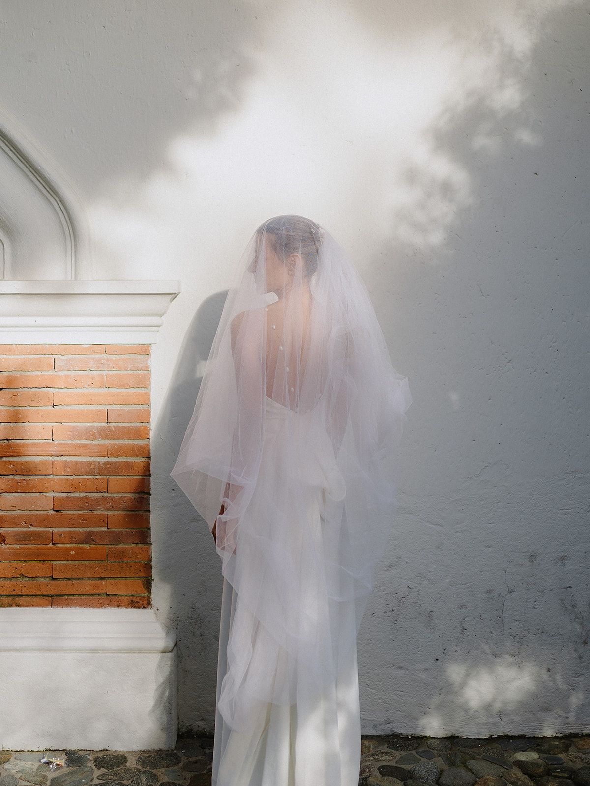 Artistic bridal portrait showing a veiled silhouette against a white and brick wall.
