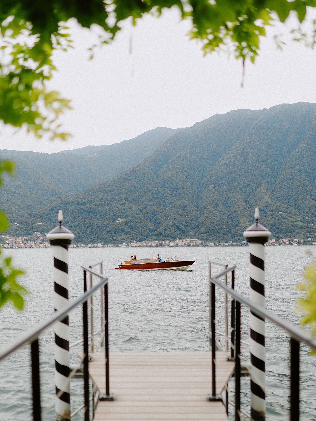 A wooden dock with striped poles frames a scenic mountain lake view with a small boat in the distance.