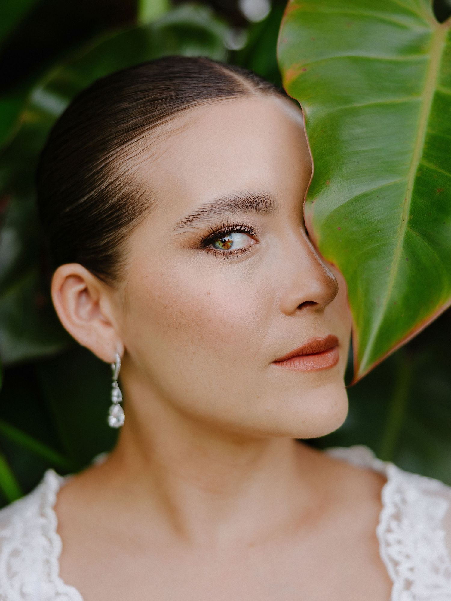 Close-up portrait with tropical leaf backdrop showing elegant bridal makeup and pearl earring.