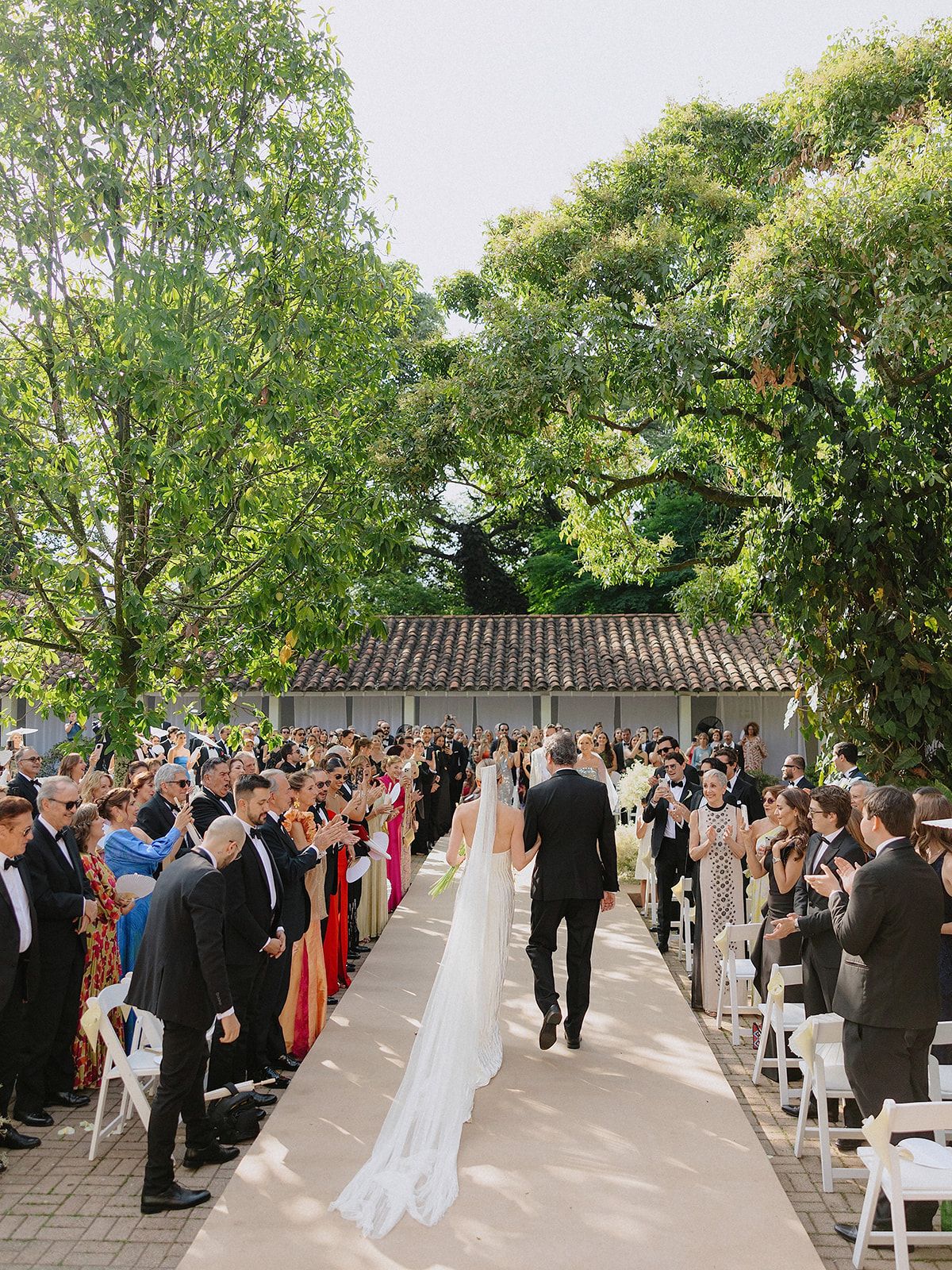 Outdoor wedding ceremony with guests lined up along a path surrounded by lush green trees and foliage.
