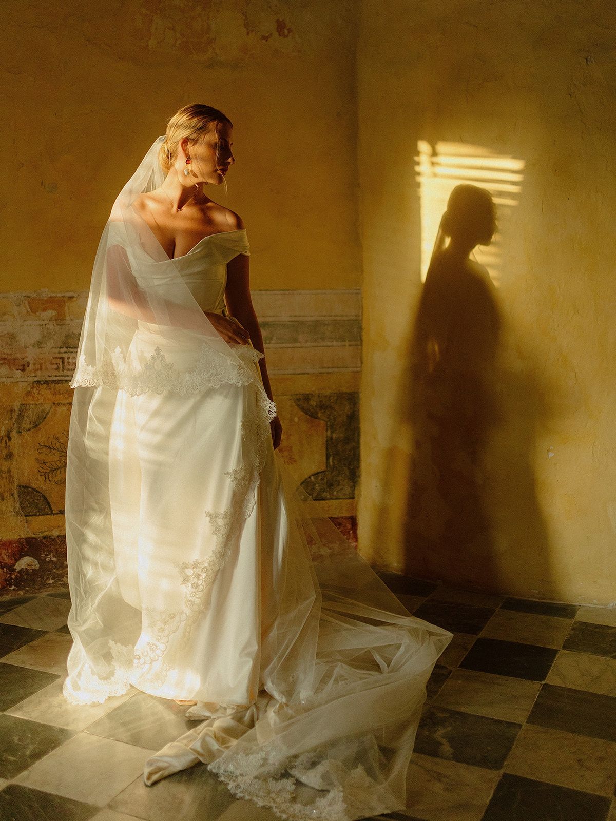 A bride in white dress casting dramatic shadows in golden sunlight on checkered floor.