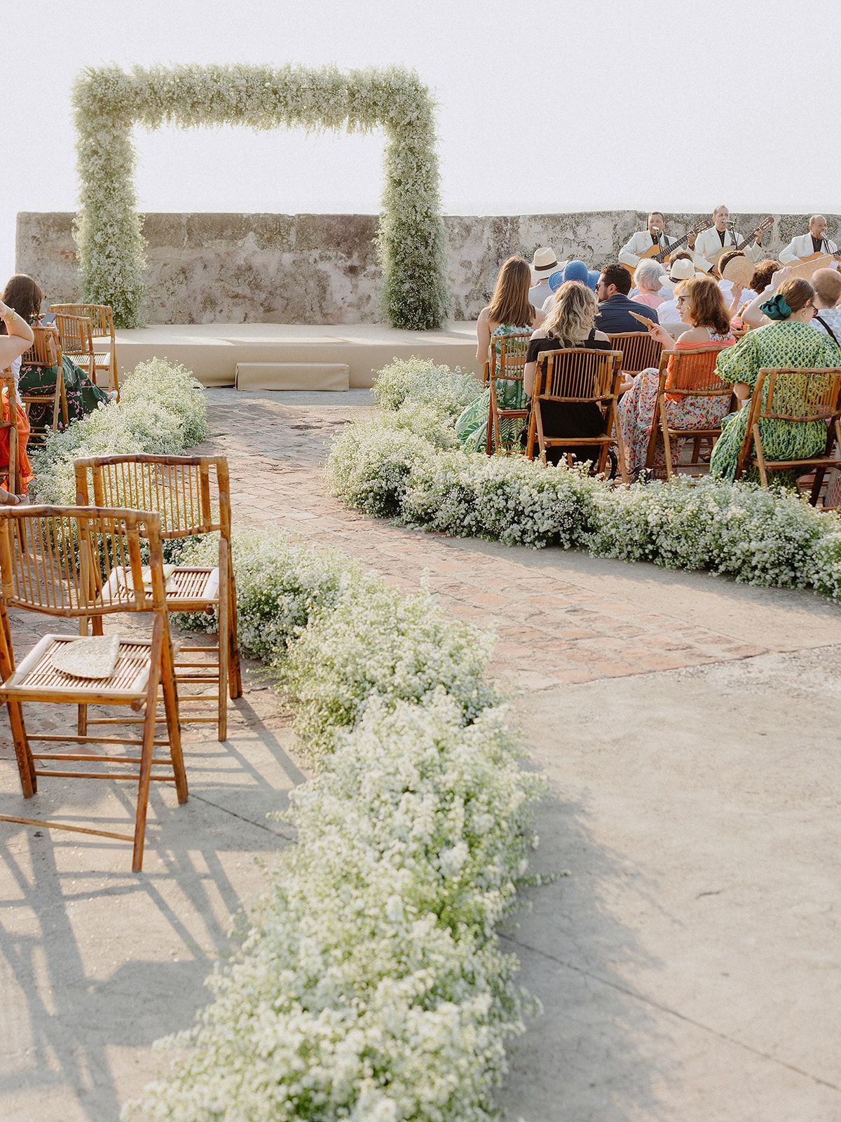Wedding ceremony setup with rows of bamboo chairs and white floral aisle decorations under an outdoor arch.