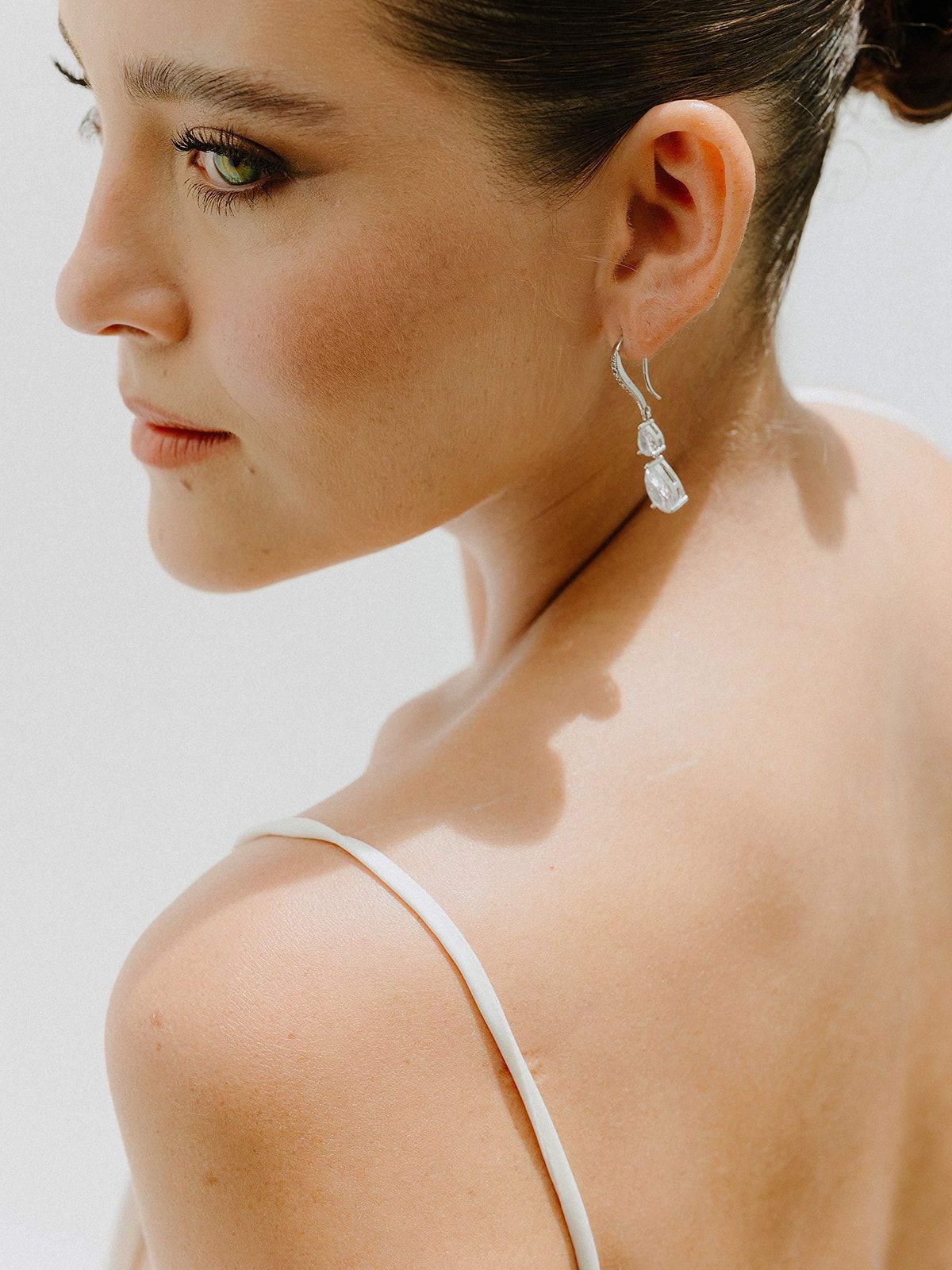 A simple studio portrait showing elegant drop earrings and a white dress strap against a light background.
