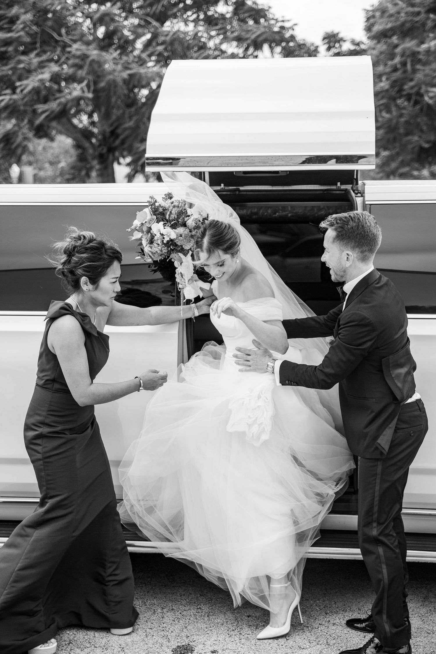 Black and white photo of a bride being helped out of a limousine by a bridesmaid and groomsman on her wedding day.