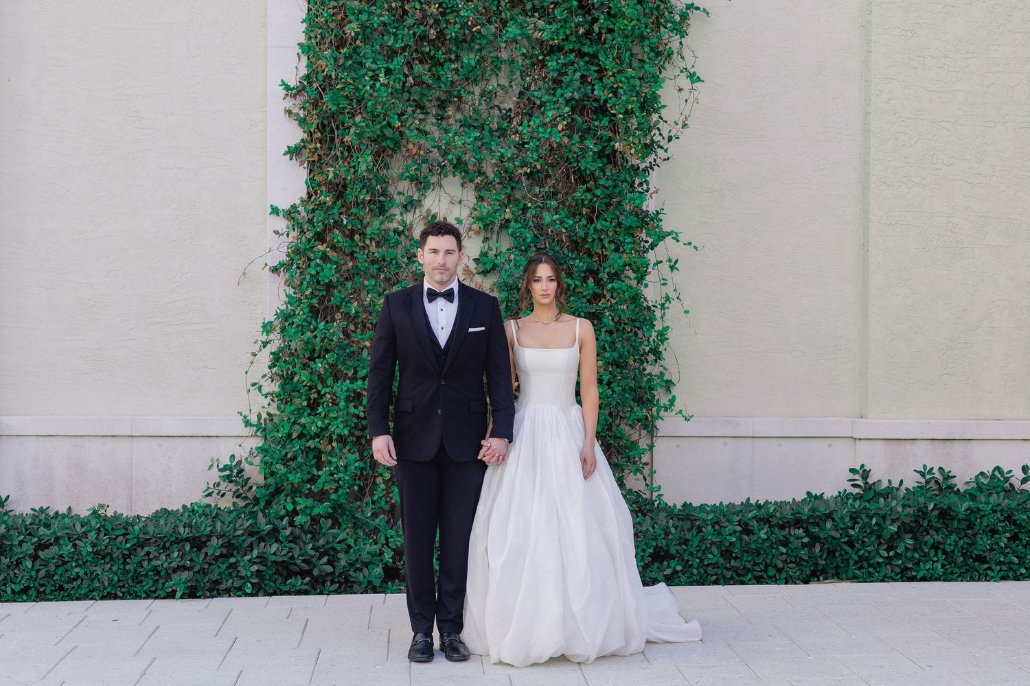 Bride and groom stand holding hands before an ivy-covered wall, she in a white gown, he in a black tuxedo.