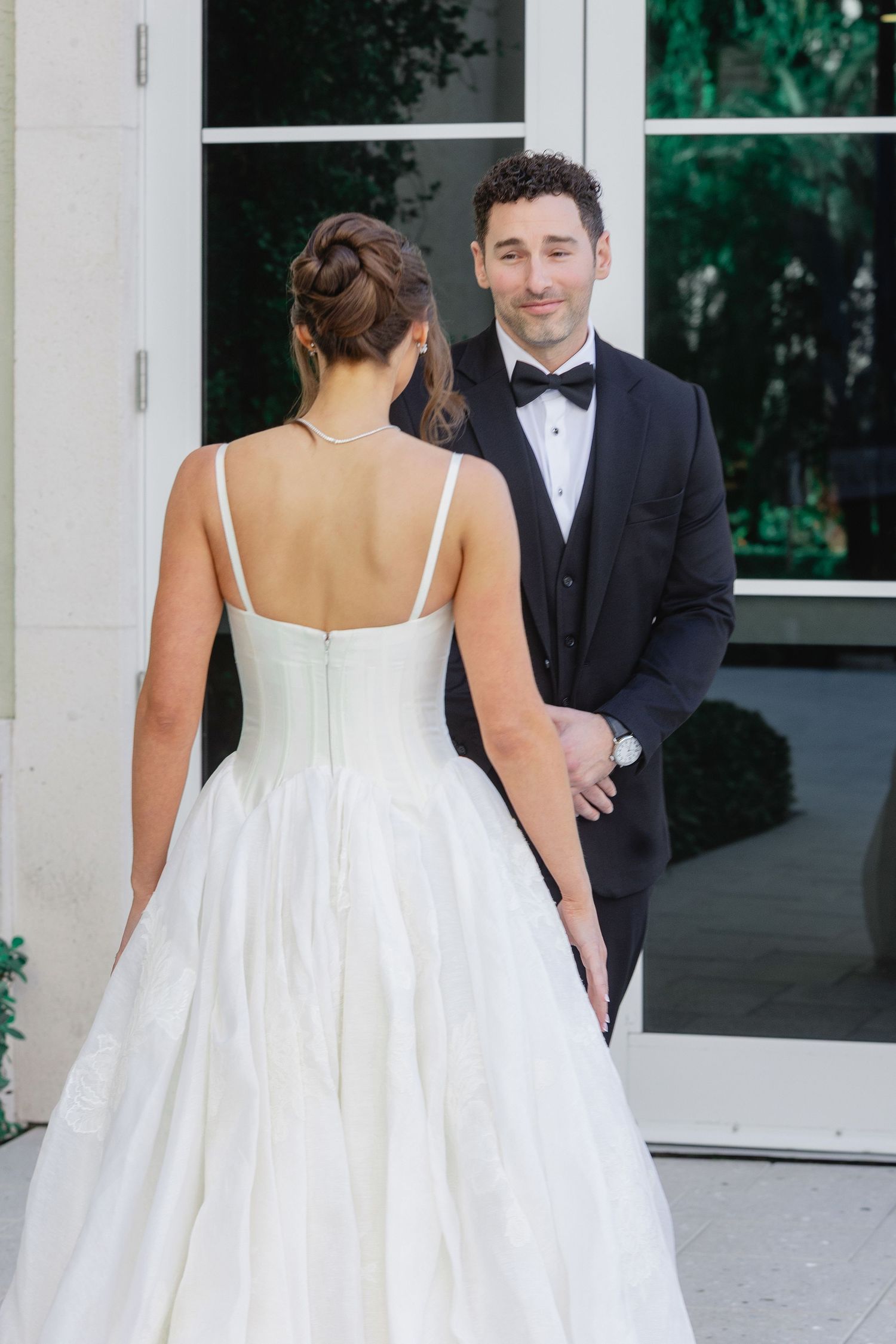 Bride in white spaghetti strap gown faces groom in black tuxedo during romantic first look at elegant venue.