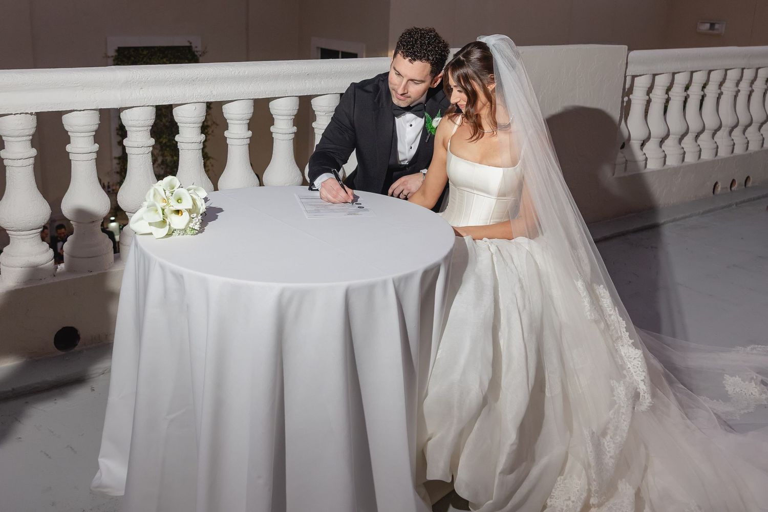 Bride and groom signing wedding documents at a white draped table, with a bouquet nearby.