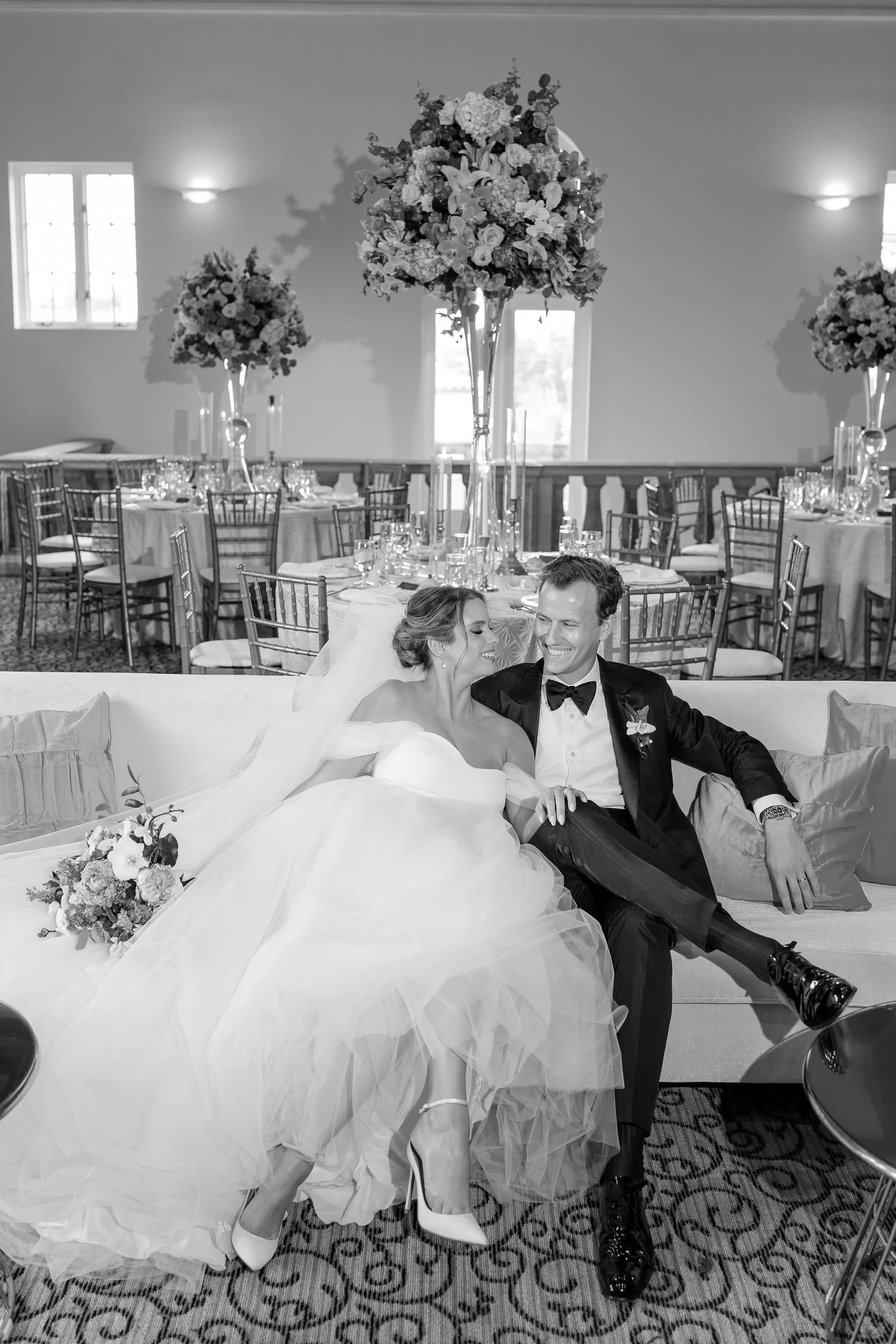 Black and white photo of a bride and groom sitting together on a sofa in an elegant wedding reception hall.