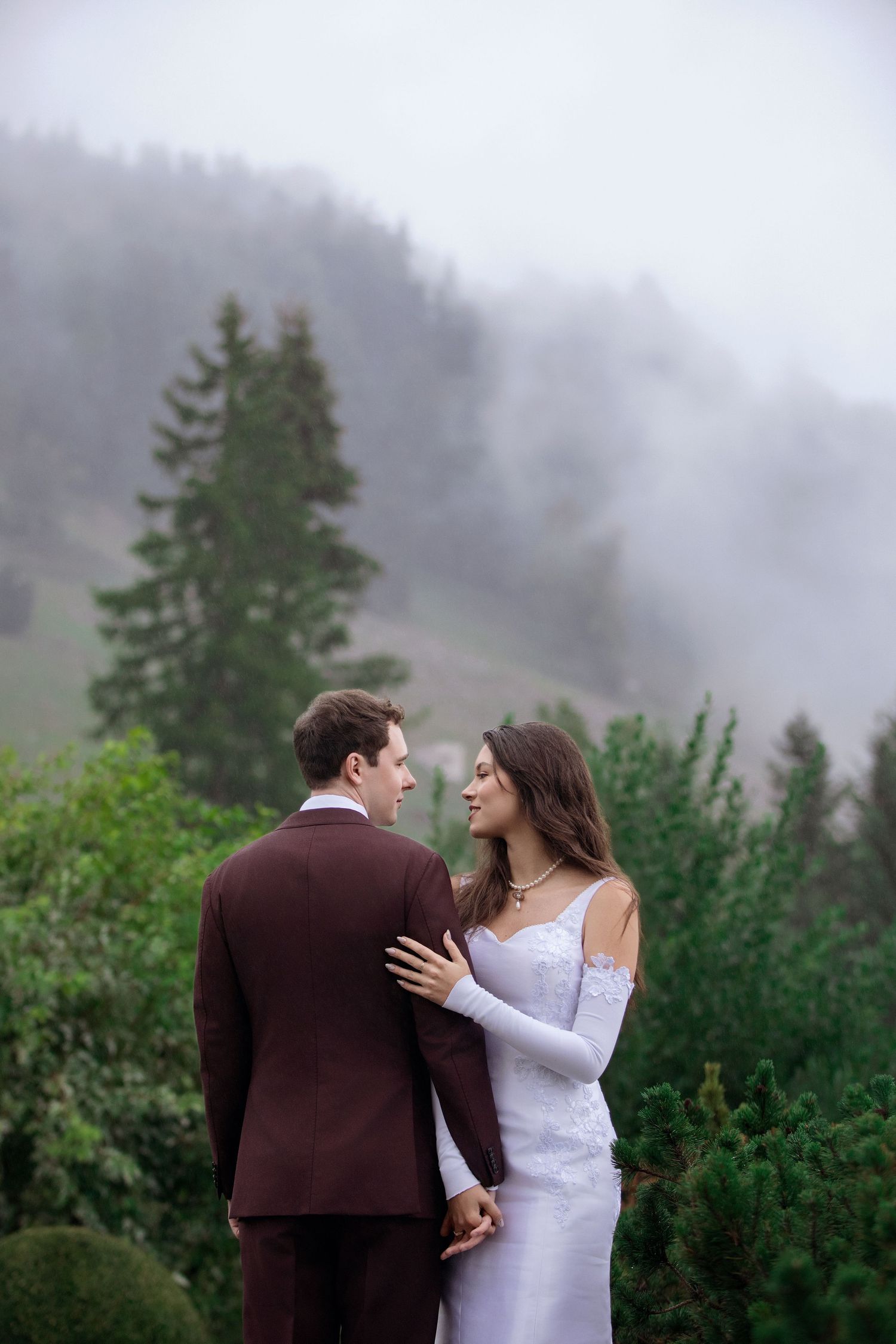 Bride and groom embrace in misty mountain forest, she wears white gown, he wears burgundy suit.