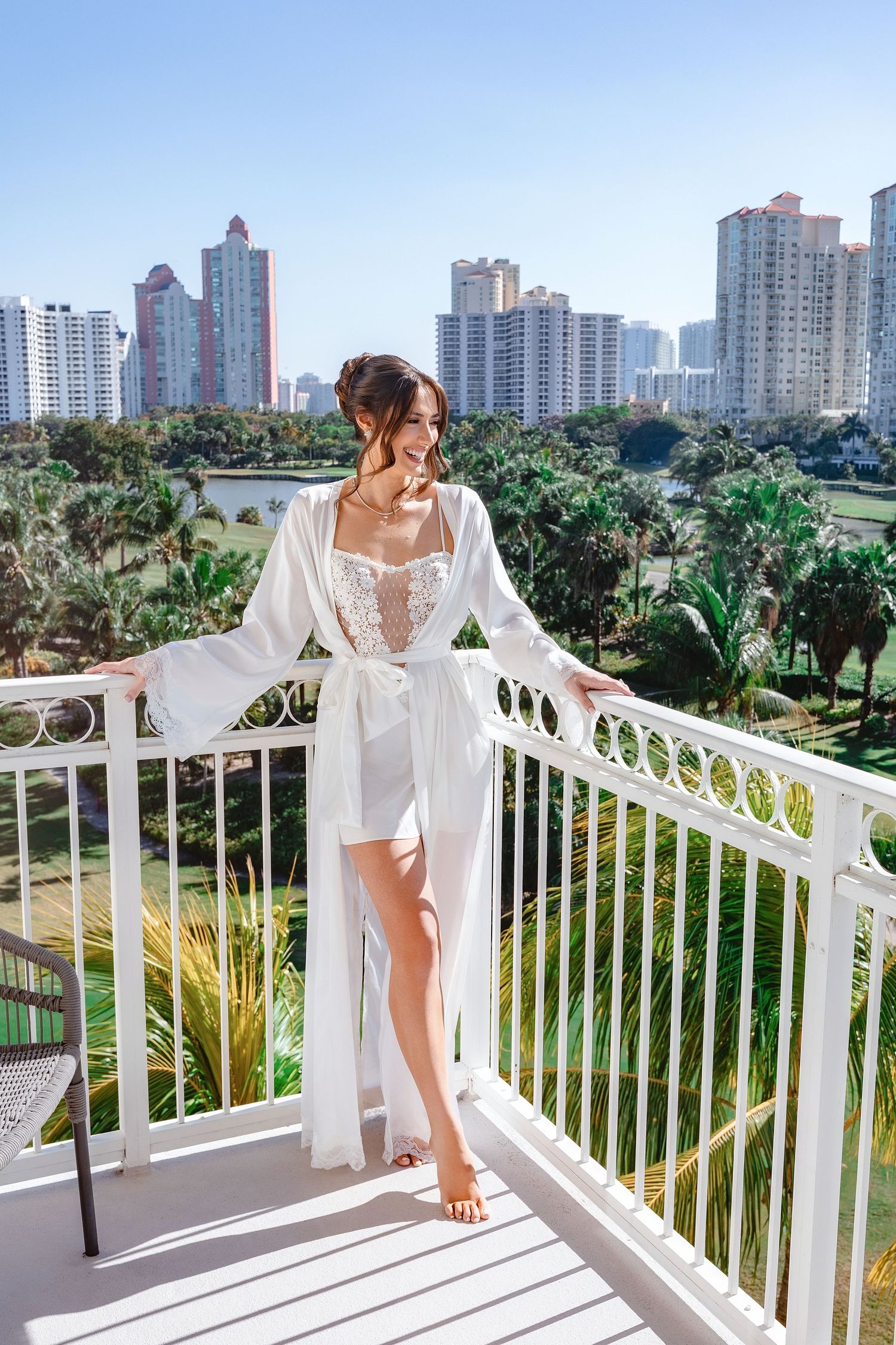 Woman in white lace bridal robe standing on hotel balcony with tropical palm trees and city skyline backdrop.