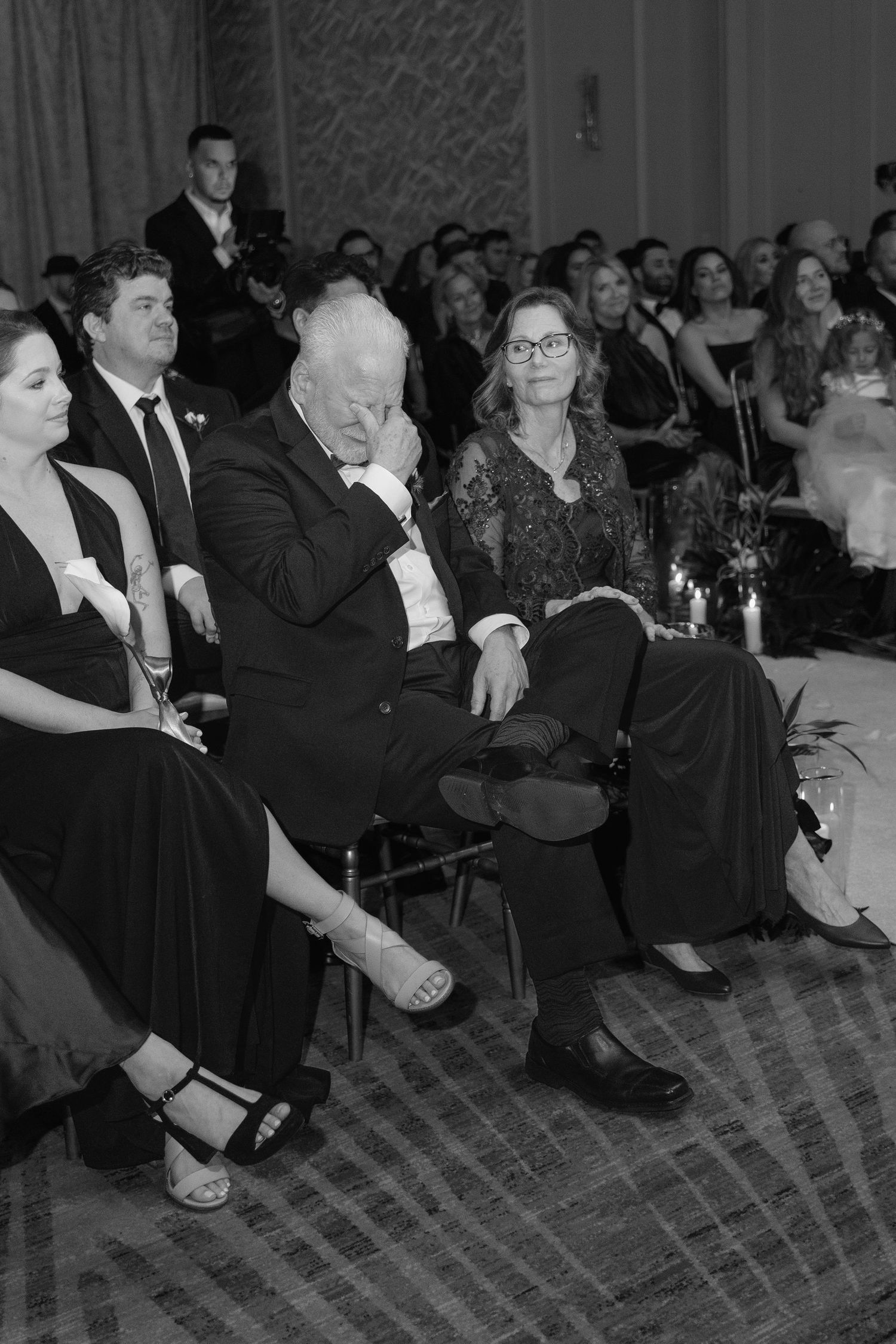 Black and white photo of emotional wedding guests seated during ceremony, man wiping tears in formal attire.