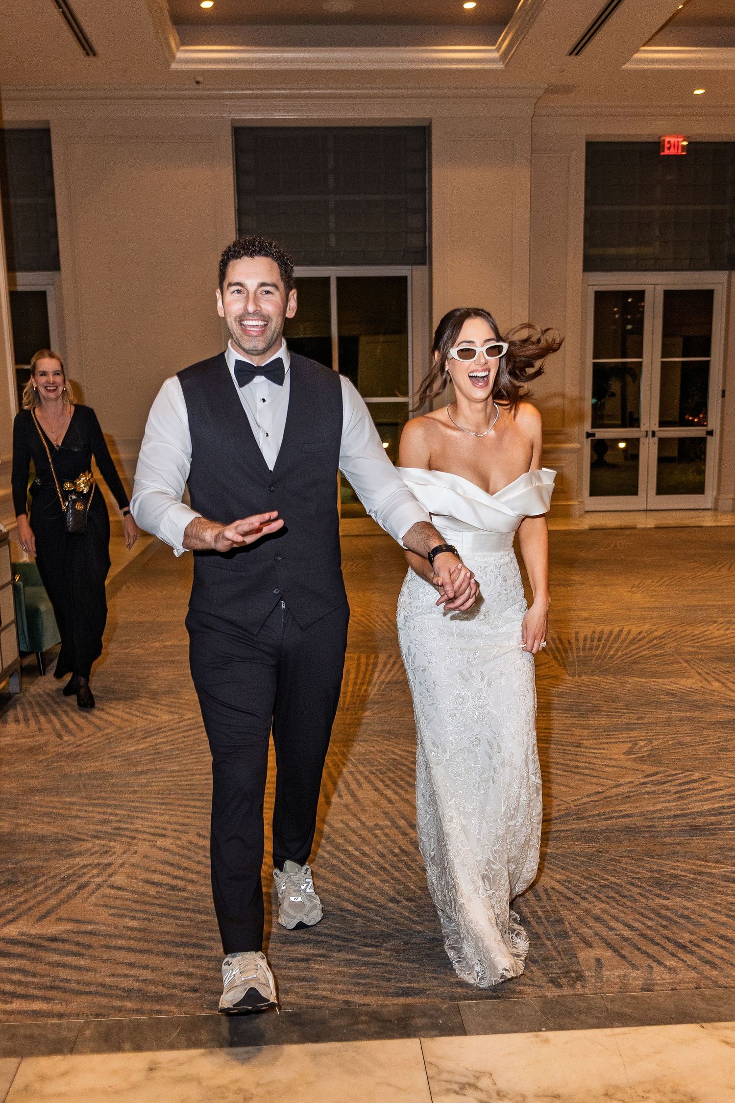 Bride in white gown and sunglasses walks with groom in tuxedo at wedding reception entrance.