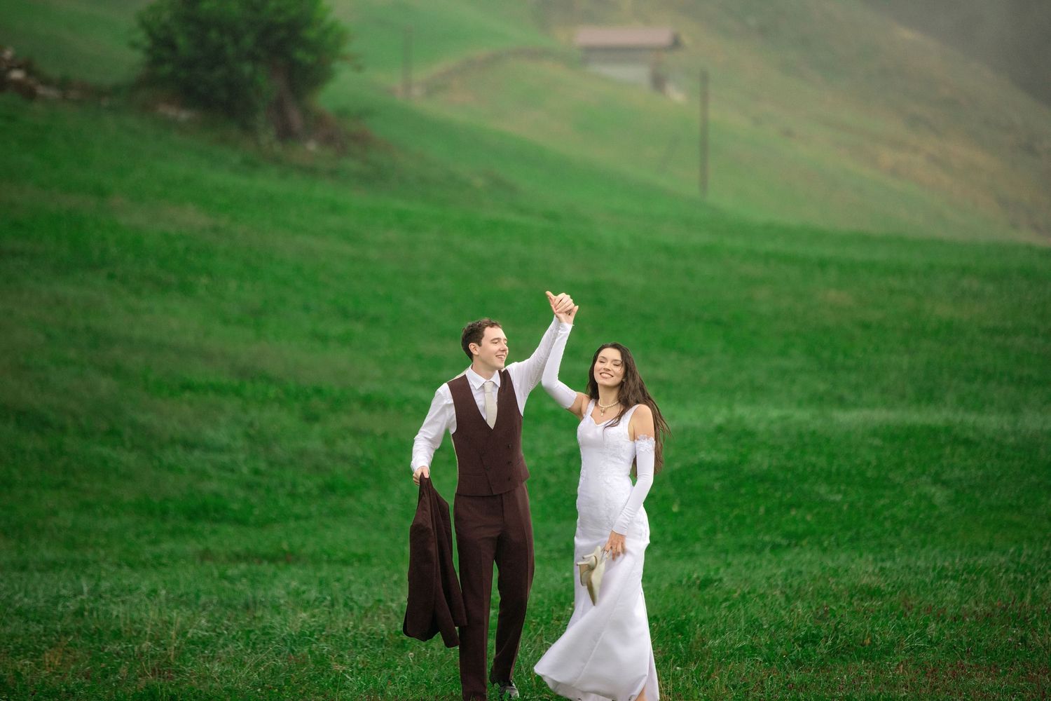 A joyful bride and groom dance in a lush green meadow, with rolling hills in the background on their wedding day.