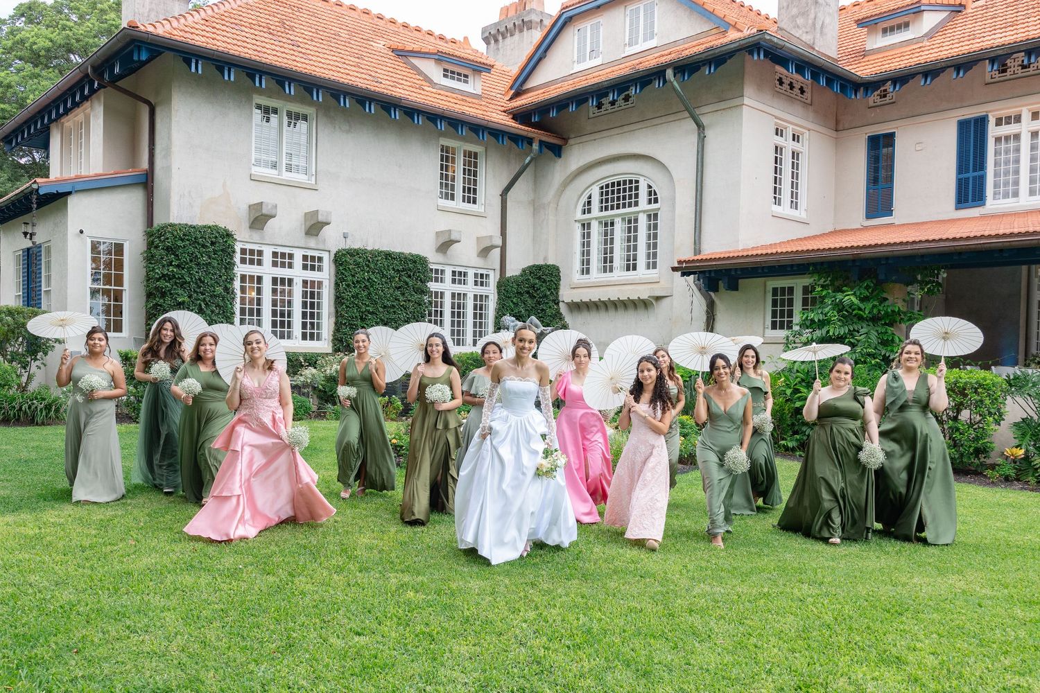 Bridal party in pink and sage green dresses poses on a manicured lawn outside an elegant white mansion.